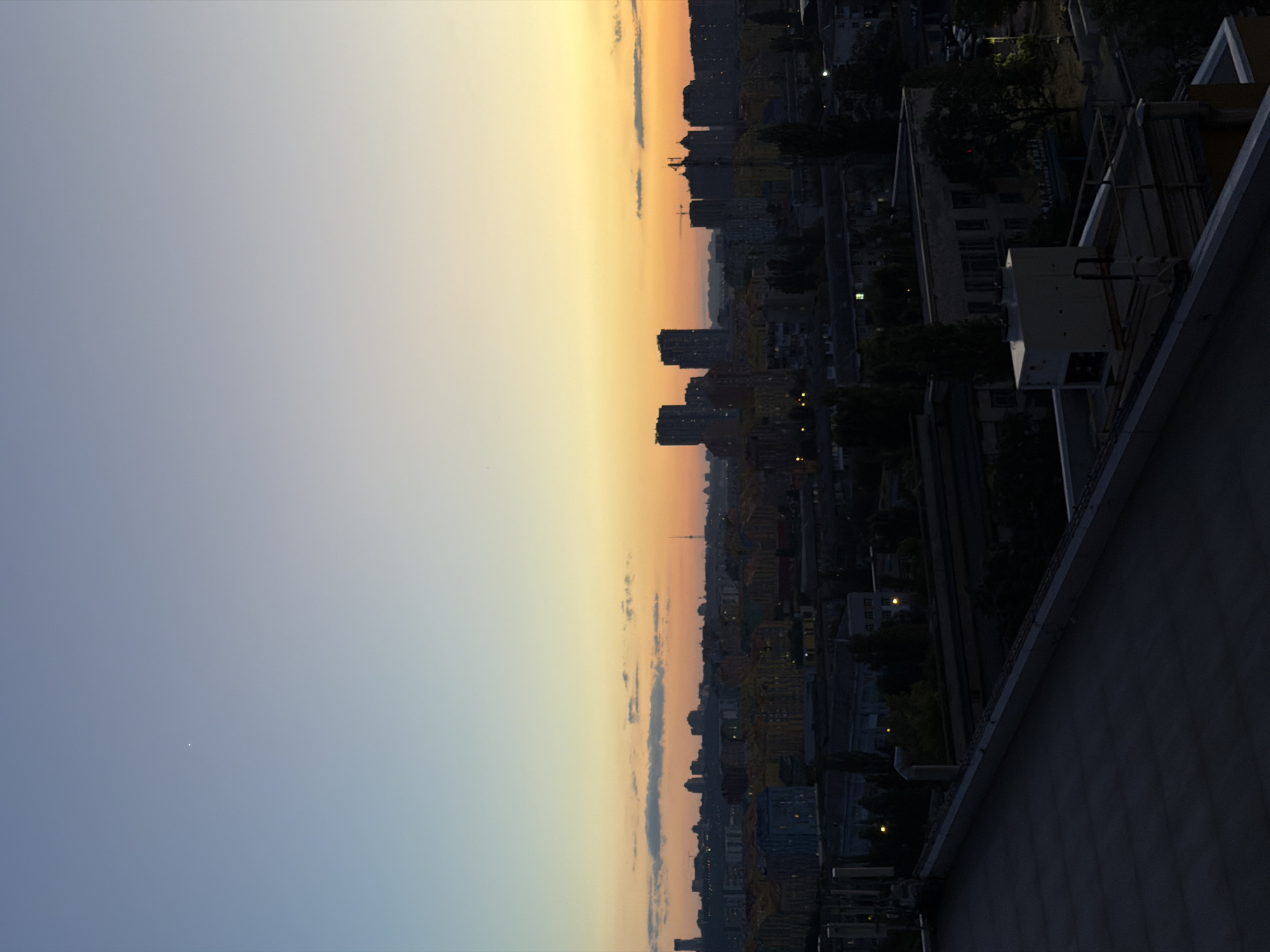 The image captures a wide-angle view of a city skyline at dusk, likely Kyiv, Ukraine, given the architectural style. The sky transitions from a pale blue at the top to a warm orange and yellow hue near the horizon, indicating the setting sun. A single, small bright point, possibly a star or a distant light, is visible in the upper left quadrant of the sky.
The cityscape below is densely populated with buildings of varying heights. In the middle ground, several modern, tall apartment buildings with dark facades are prominent, interspersed with lower-rise structures and what appear to be industrial or commercial buildings. Some windows in these buildings are illuminated, suggesting activity within.
In the foreground, the dark, flat roof of a building dominates the lower portion of the image. A large, white, rectangular air conditioning unit is situated on this roof, along with some metal railings and other rooftop equipment. The overall scene is characterized by a sense of urban sprawl and the transition from daylight to evening, with artificial lights beginning to punctuate the darkening landscape. There are no people or visible actions depicted in the image.