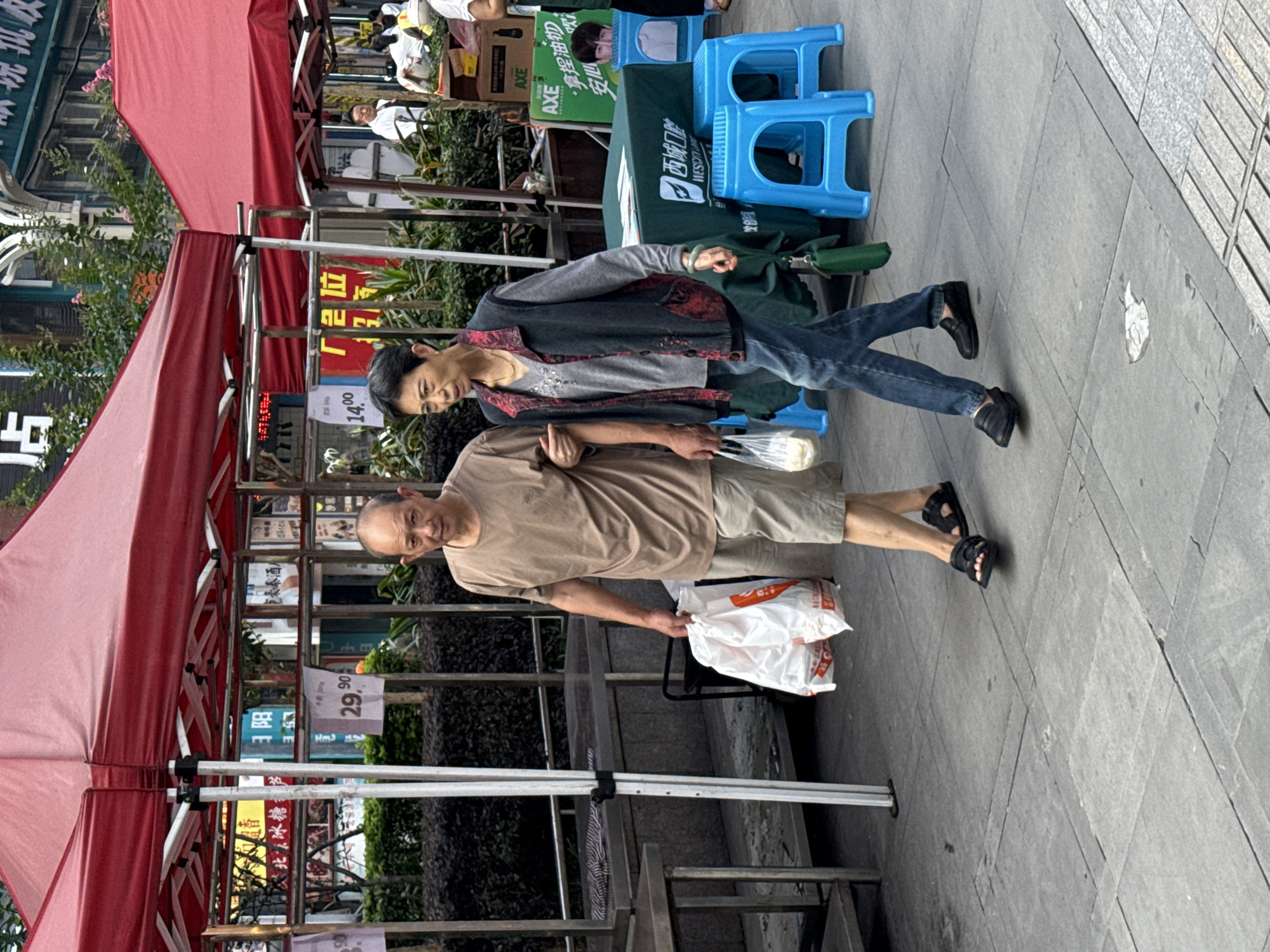 An elderly Asian couple walks together on a sidewalk lined with outdoor market stalls in Jiangjin, China. The man, on the left, wears a light brown t-shirt and khaki shorts, and carries a large white plastic bag in his right hand. His left arm is around the woman's shoulders. The woman, on the right, is dressed in a dark grey vest with embroidered red and black floral patterns over a light grey v-neck shirt, blue jeans, and black slip-on shoes. She carries a clear plastic bag in her right hand and her left arm is linked with the man's. They are walking from right to left in the frame. Behind them, red-canopied market stalls are visible, displaying various goods. Several people can be seen in the background browsing or working at the stalls. The ground is paved with square gray tiles, and some of the stalls have tables and blue plastic stools set up. The overall atmosphere suggests a busy market day.
