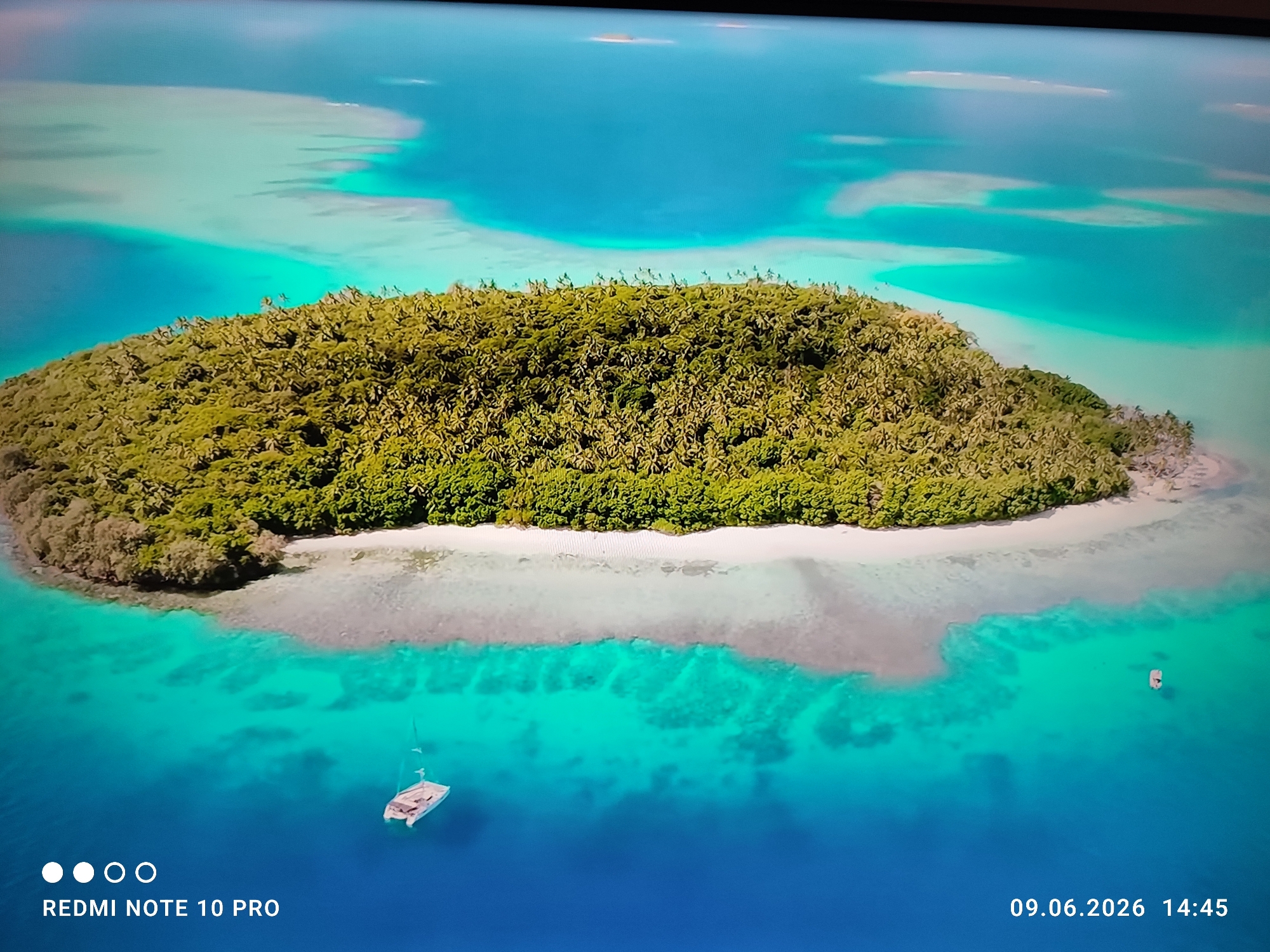 An aerial drone photograph captures a lush, densely vegetated island surrounded by vibrant turquoise and deep blue ocean waters. The island, covered in palm trees and other greenery, is fringed with a white sandy beach. A small sailboat with a white hull and mast is anchored in the clear, shallow water near the shore. In the distance, a few small, sandy atolls are visible in the expansive ocean. The image includes a date stamp of "09.06.2026 14:45" and "REDMI NOTE 10 PRO" text at the bottom. The lighting suggests a bright, sunny day with the sun high in the sky, casting minimal shadows. The scene is serene and idyllic, evoking a tropical paradise.