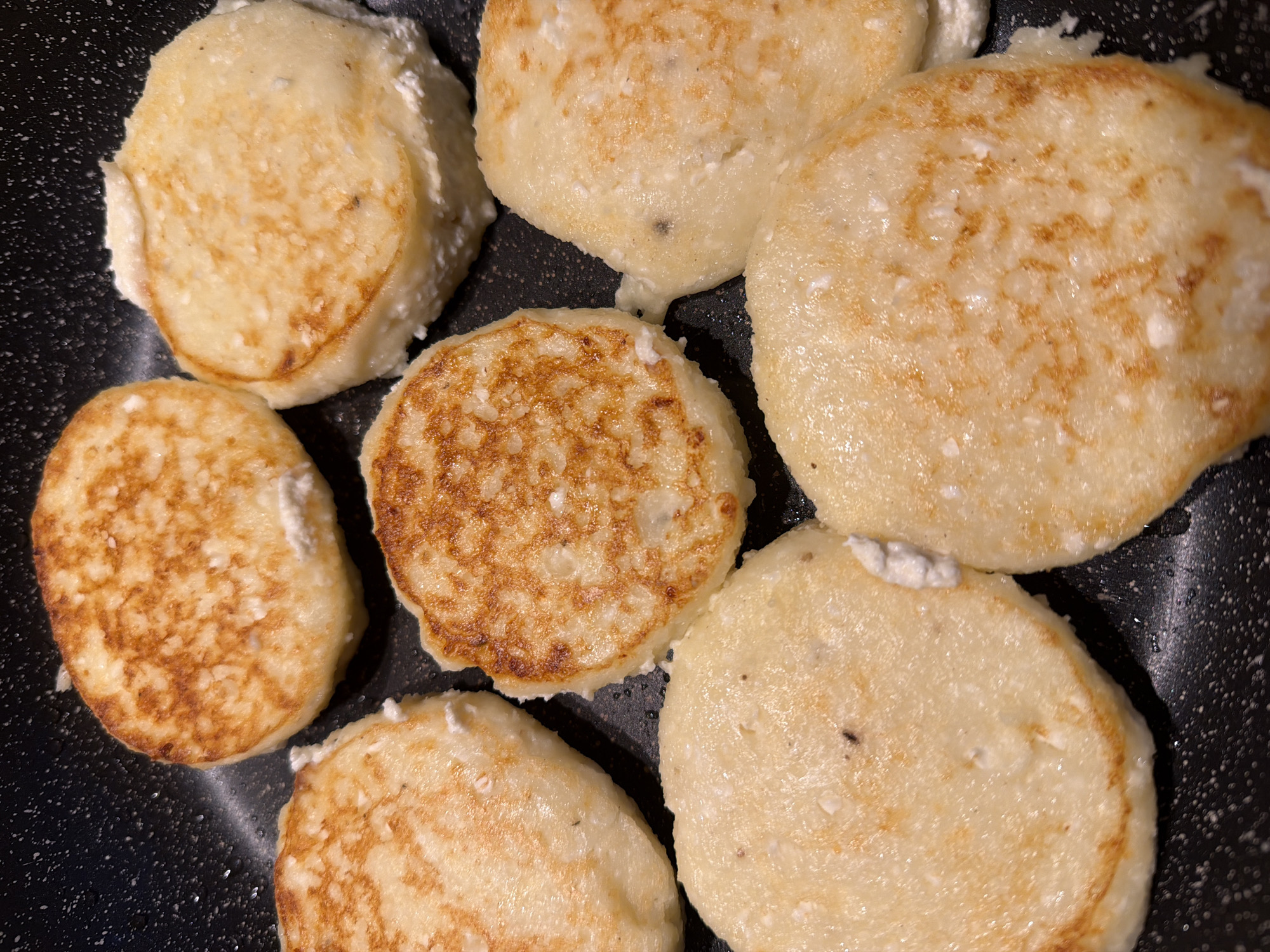 The image shows seven syrniki (a traditional Russian and Ukrainian cheese pancake) in a dark frying pan. They vary in doneness, with one being darker and more cooked than the others. Some have small pieces of uncooked cheese visible. The frying pan has a speckled appearance, potentially indicating a non-stick surface. The location is not discernible from the image.