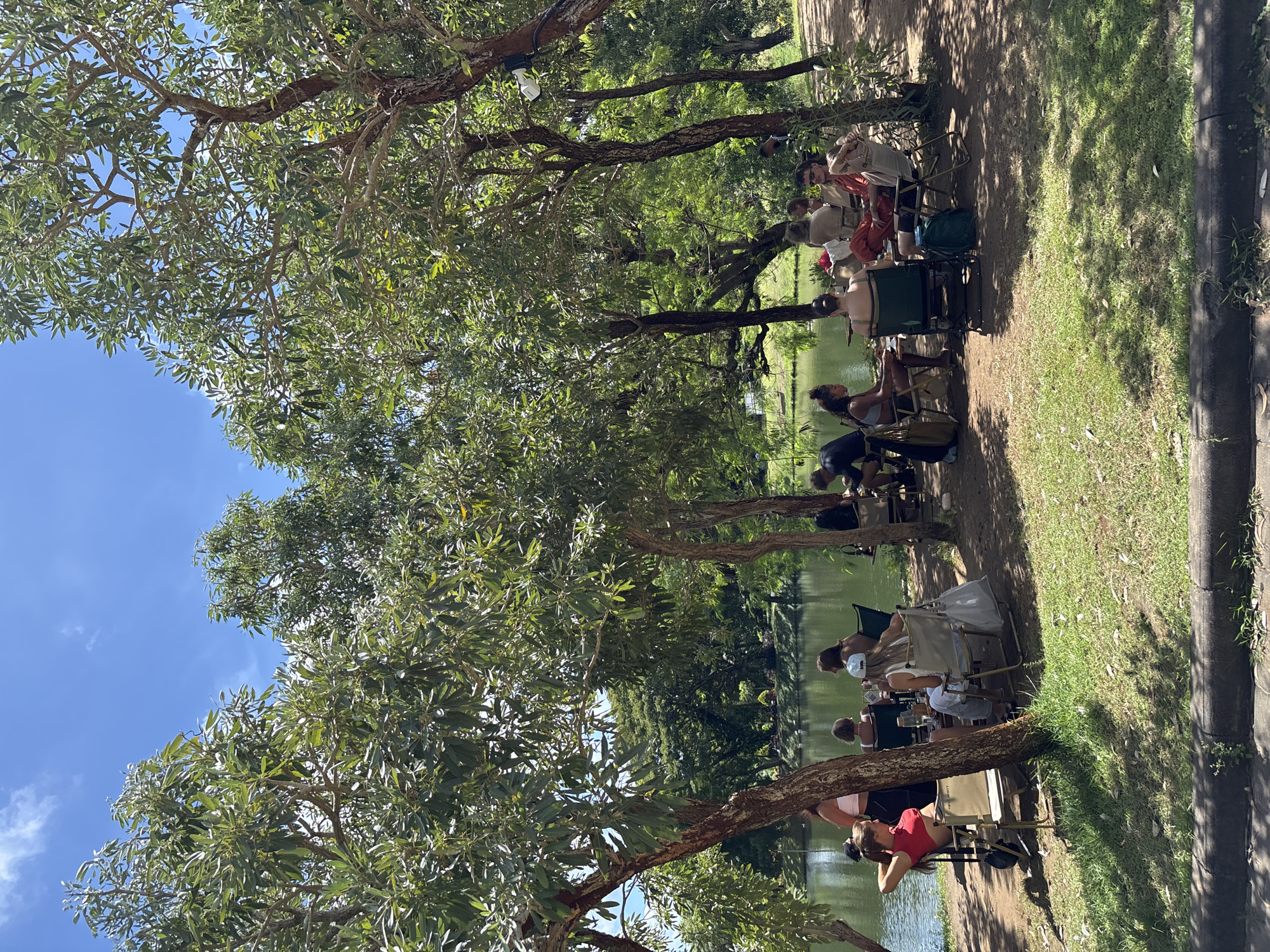 A group of people sits in chairs along a grassy area next to a body of water, possibly a lake or pond. They are shaded by trees with dense foliage. The location appears to be outdoors, likely in a park or recreation area within the city of Kuta Selatan, Indonesia. The sky is blue, and the weather seems sunny.