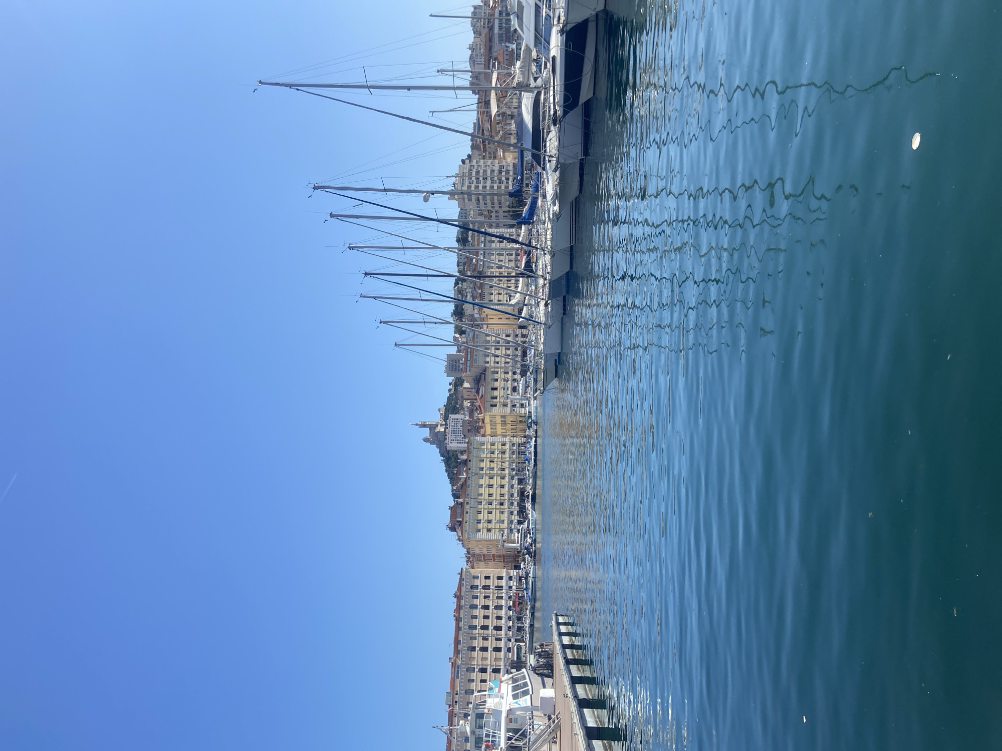 The image depicts a bright, sunny day in a harbor filled with sailboats. The foreground is dominated by calm, blue water with reflections of the sky and the sailboats. Along the left side of the frame, a wooden dock and the side of a white boat are visible. In the background, a row of sailboats with tall masts lines the harbor. Beyond the sailboats, a city skyline is visible, characterized by multi-story buildings with terracotta roofs. Atop a hill in the distance, a prominent white building with a dome, likely a basilica or church, stands out against the clear blue sky. The overall scene conveys a sense of a peaceful, well-maintained harbor in a coastal city.