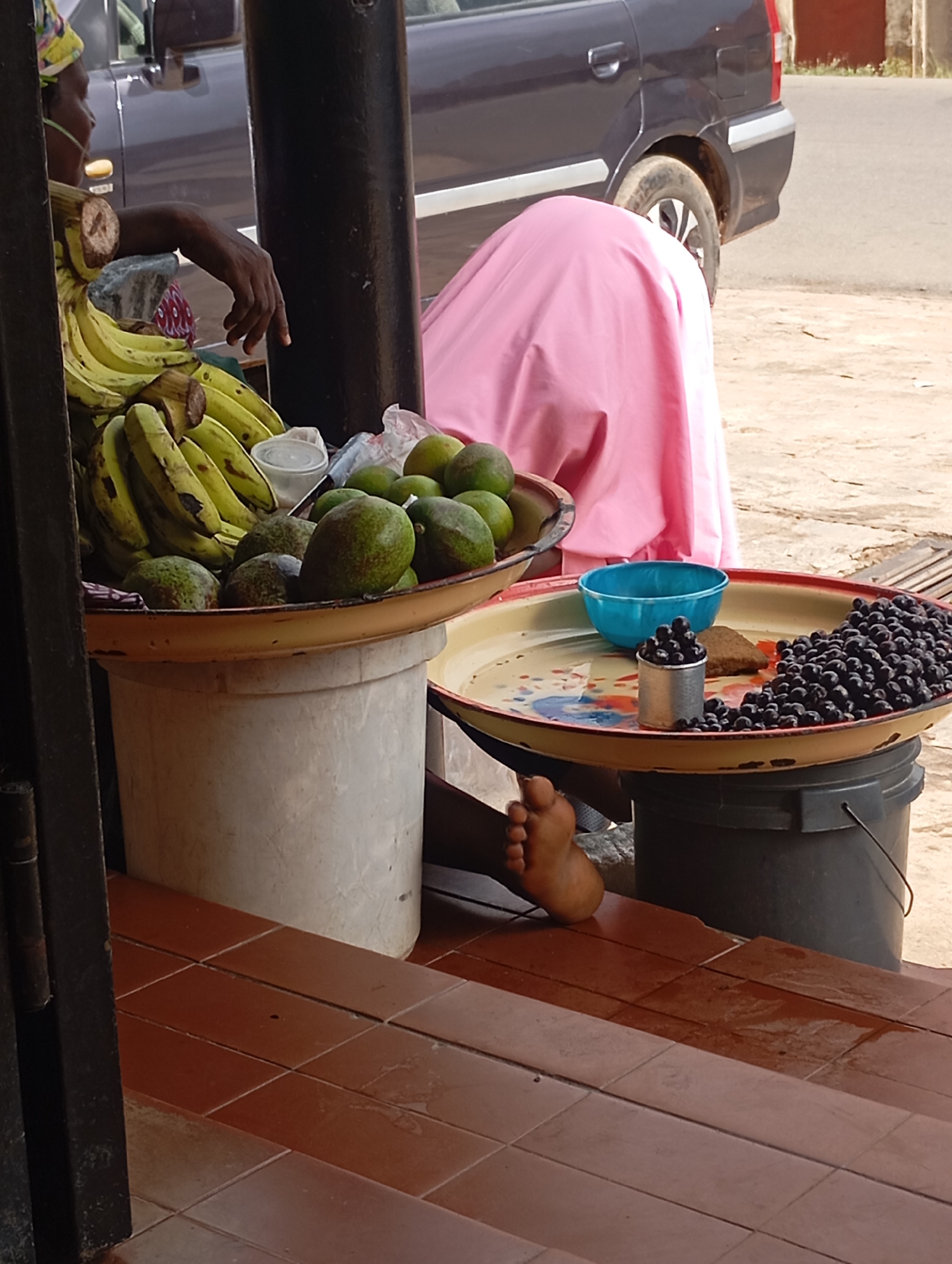 The image shows a person sitting at what appears to be a market stall in Jos, Nigeria. The person, wearing a yellow and green headwrap, is partially visible, with their arm resting on a collection of ripe bananas. In front of the bananas, on a large, shallow tray, are several avocados. The tray is placed on top of a large white bucket.
Another tray, resting on a dark gray bucket, holds a blue bowl, a small piece of baked goods, and a pile of dark berries, possibly blueberries or blackcurrants, contained in a small silver tin. The background shows a dark-colored vehicle and a woman covered in a pink cloth, also appearing to be at the market. A bare foot is visible near the edge of the foreground, resting on red tiles. The scene suggests a sunny day, likely during market hours, with goods being sold. There are no visible text elements in the image.