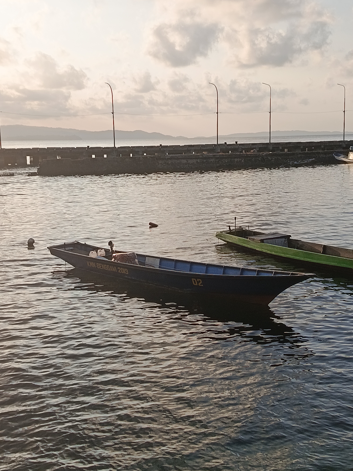 This image depicts a serene waterfront scene in Mansalean, Indonesia, during what appears to be either sunrise or sunset. The foreground is dominated by calm, rippling water reflecting the soft light. Two small boats are visible. The closest boat is a dark blue vessel with the text "KMM-BENGGANI 2019" and the number "02" clearly marked on its side. Inside this boat are two white jerrycans and some covered cargo. A second, longer boat, painted green with a dark interior, is moored slightly behind and to the right of the blue boat.
In the mid-ground, a concrete breakwater stretches across the scene. The breakwater is lined with multiple street lamps on tall poles, suggesting this is a developed waterfront area. Beyond the breakwater, a distant shoreline with rolling hills is visible, partially obscured by a light haze. The sky is filled with soft, scattered clouds, bathed in the warm, muted light of either dawn or dusk. The overall atmosphere is tranquil and peaceful.