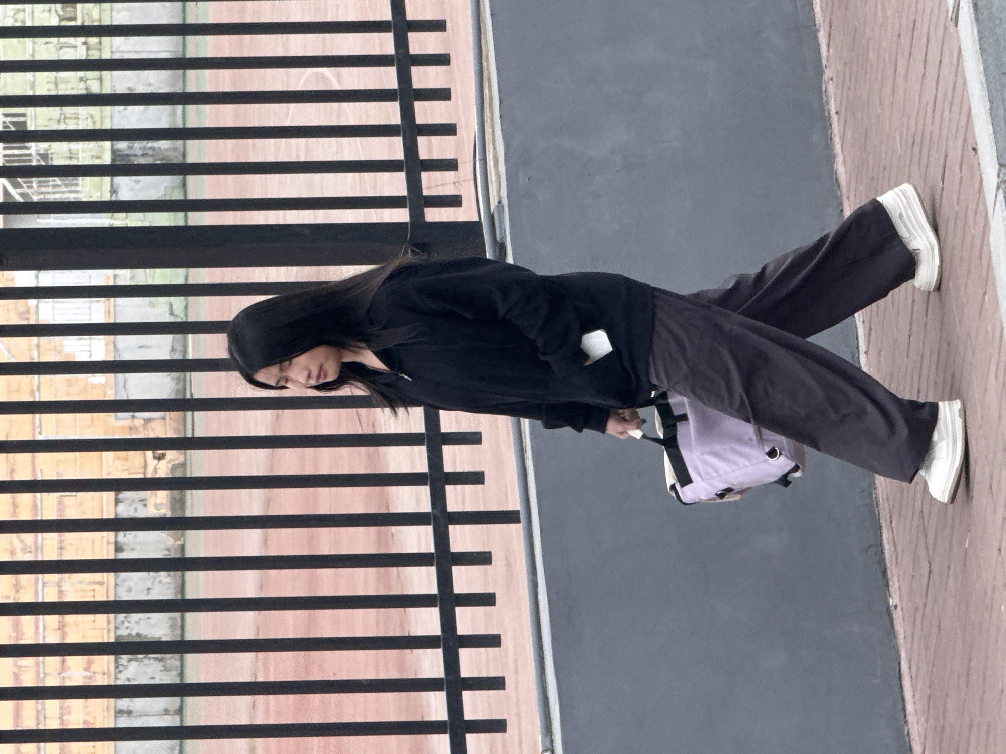 A person with long, dark hair walks outdoors in what appears to be an urban or semi-urban setting. They are dressed in a dark (black or very dark grey) long-sleeved hooded top and matching wide-leg or straight-leg pants. White sneakers with distinctive soles complete their casual attire. The person carries a light-colored, possibly light purple or off-white, shoulder bag with black straps in their right hand, holding it by their side. Their head is slightly bowed, indicating focus as they walk.
The ground they are traversing is a reddish-brown paved or tiled surface. Adjacent to this, a darker grey surface, perhaps asphalt or concrete, is visible. A prominent feature of the background is a dark metal fence composed of numerous horizontal bars, running across the scene behind the person. Through the gaps in the fence, blurred details of buildings or structures are discernible, displaying light-colored brickwork or concrete facades. The lighting is soft and even, suggesting it is daytime, possibly under an overcast sky, as there are no harsh shadows. No visible text is present in the image.