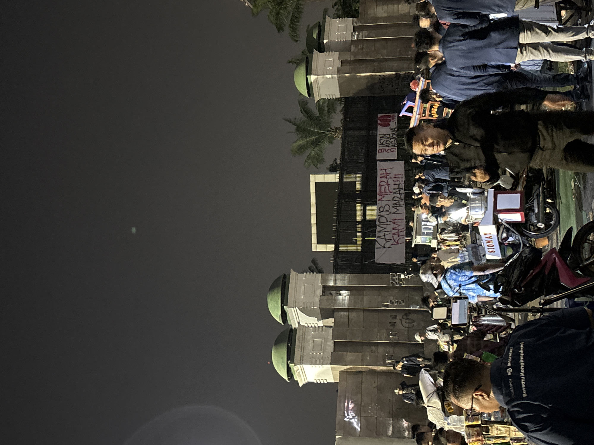 The image depicts an outdoor night scene in Central Jakarta, Indonesia, possibly near a government building or historical landmark, given the architectural style of the structure in the background featuring two domed towers. A crowd of people, mostly young adults, are gathered, suggesting an event or protest. Several banners are visible; one reads "KAMPUS MERAH KAMI MARAH!!!" which translates to "RED CAMPUS WE ARE ANGRY!!!" Another banner reads "BIKIN GADUH NASIONAL" meaning "CREATE NATIONAL CHAOS." The atmosphere appears to be tense. In the foreground, a man in a black jacket is gesturing with his hand, while other individuals are seen in conversation or observing the surroundings. Various market stalls or food vendors are set up, indicated by the presence of carts and goods. The lighting is dim, with artificial lights illuminating parts of the scene, and a slight lens flare is noticeable.