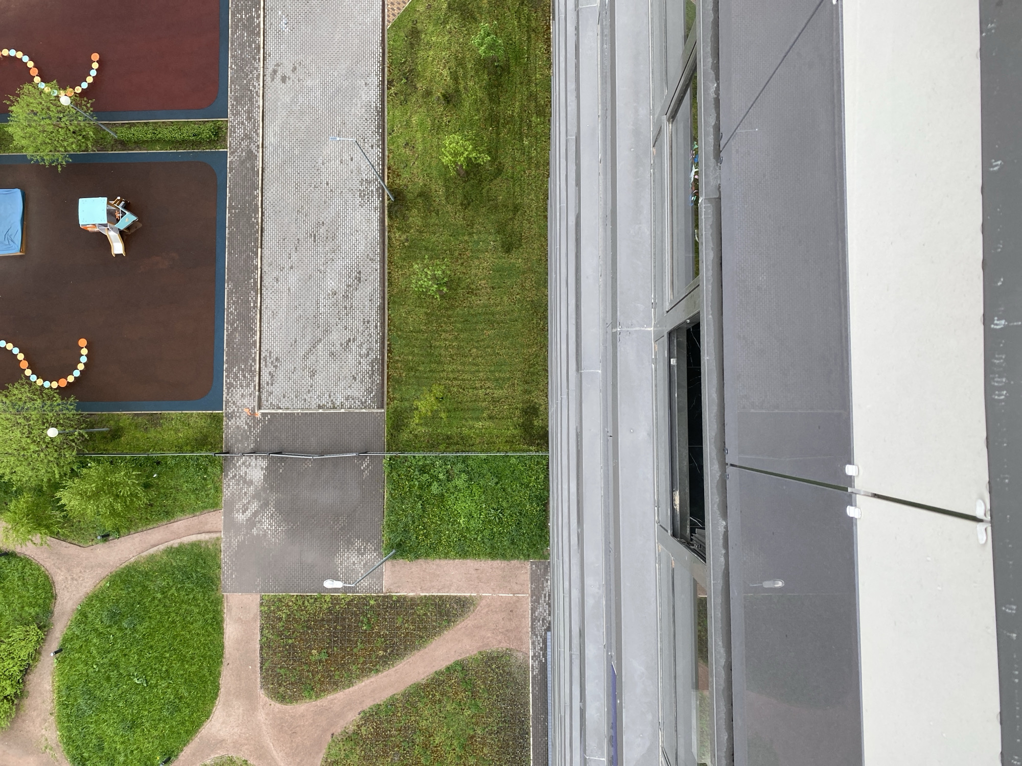 The image shows an aerial view of a scene consisting of a playground, walkways, grassy areas, and a building. The playground is located on the left side of the image and includes a brown soft surface, playground equipment, and colorful ball decorations. Adjacent to the playground is a walkway made of gray paving stones. To the right of the walkway is a strip of green grass. Below the playground and walkway, there is a green space with walking paths. On the right side of the image is a building with gray siding and windows. The image appears to be taken from a high vantage point, likely from a higher floor of the building.