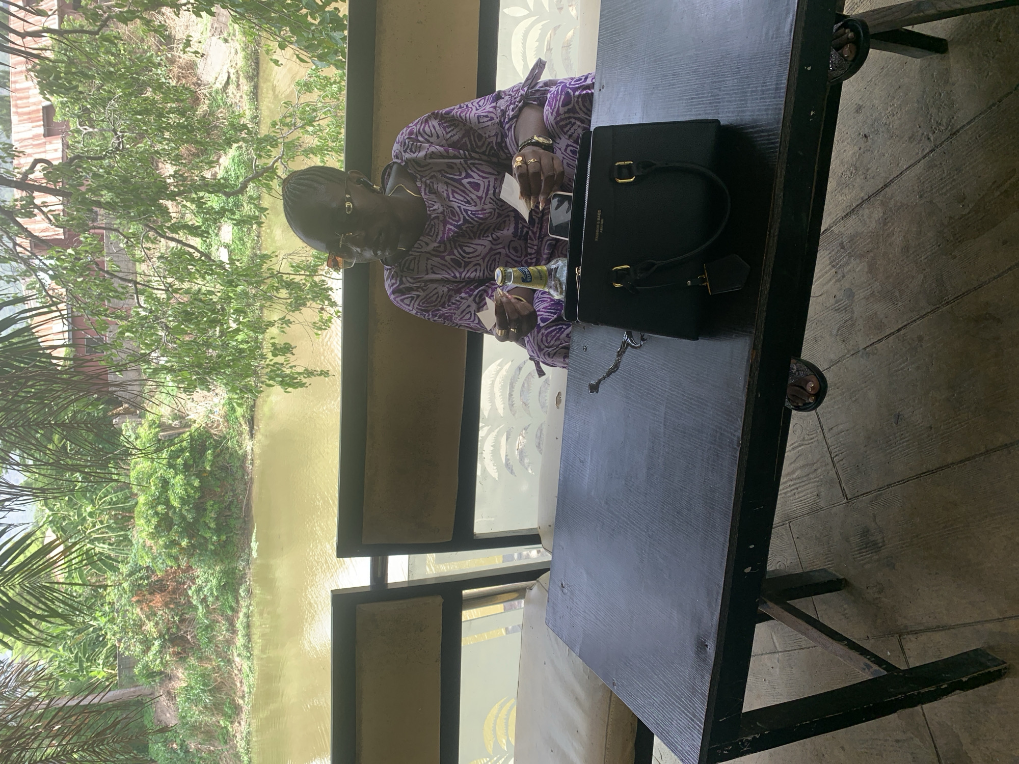 An eye-level, rotated outdoor shot shows an older woman with dark hair in Adankolo, Nigeria, seated at a black wooden table near a body of water. The woman wears sunglasses, gold jewelry and a purple patterned shirt. She is using a toothpick. On the table are a bottle of beer and a black purse with gold hardware. A view of the river and green vegetation are visible through the framed glass panels behind her. The ground appears to be concrete.