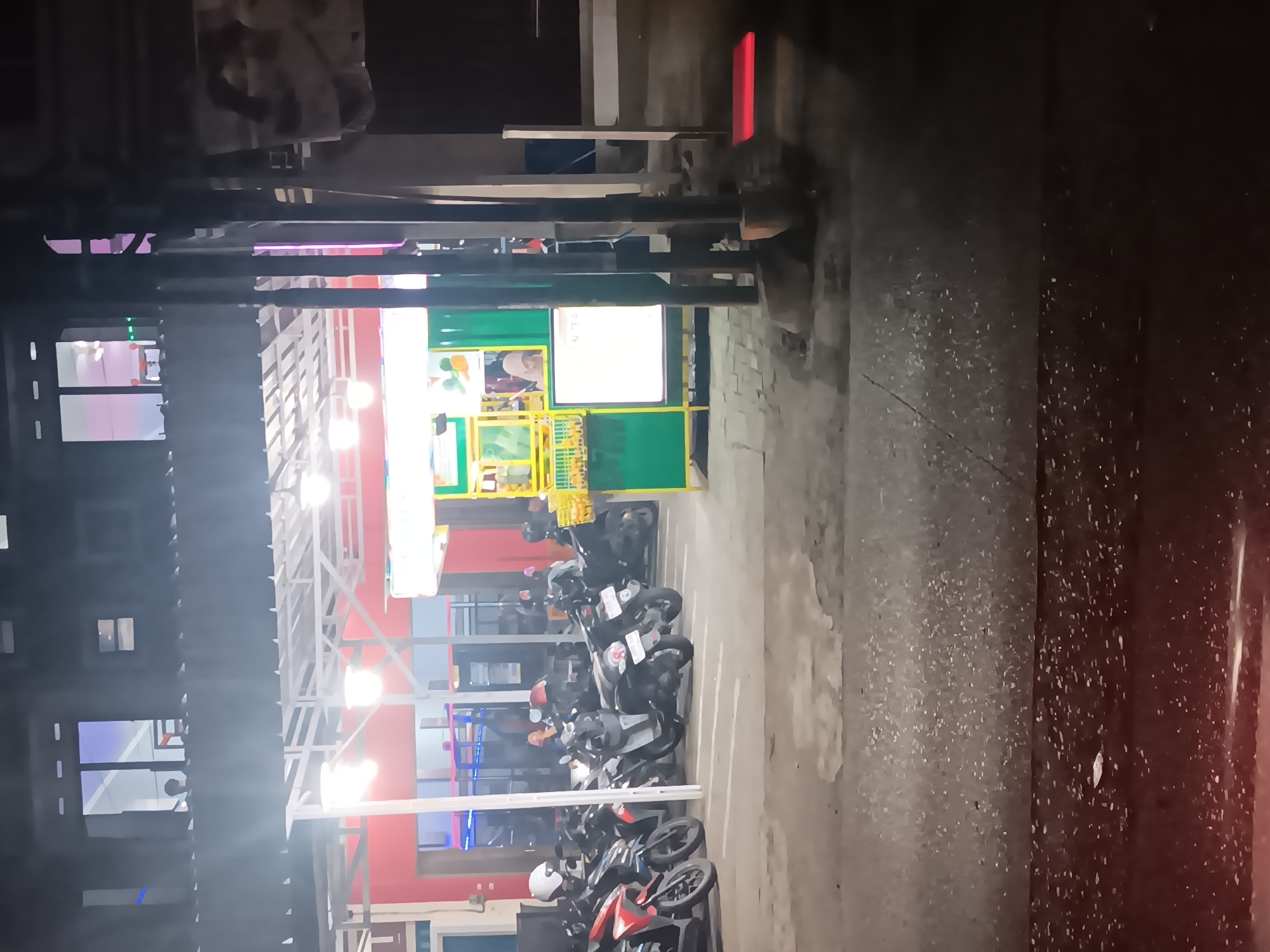 The image shows a nighttime scene of a street in Batursari, Indonesia, with a row of parked motorcycles. The motorcycles are arranged in parking spots in front of a building with a red facade and a white awning. The building appears to be a shop or restaurant, with lights illuminating the interior and a green food stall in front. A few people are visible inside the building and near the food stall. The street in front of the building is wet, suggesting recent rain or dampness, and there are reflections of lights on the surface. A lamppost and some vertical metal structures are also present in the foreground. The overall atmosphere is one of a bustling urban environment at night.