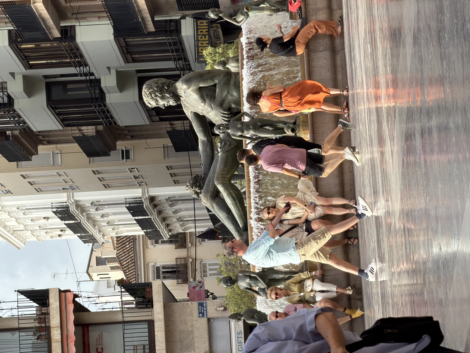 The image depicts a public plaza in an urban setting, likely in Europe given the architecture and the presence of a statue. In the foreground, a man in a light blue short-sleeved shirt and khaki shorts is walking across a wet, reflective ground. He is wearing white athletic shoes with black stripes. Behind him, several people are seated on the edge of a fountain.
The fountain itself is a prominent feature, featuring several bronze sculptures. A large male figure reclines at the top, with a female figure seated below him, holding a jug. Other smaller figures are also integrated into the fountain's design, with water cascading around them.
In the background, buildings with classical architectural elements, balconies, and shuttered windows line the plaza. One building displays signage, including "PLACA DE LA MARE DE DEU DE LA PAU" and what appears to be a store name. The sky is overcast. The scene captures a moment of everyday life in a city square, with people enjoying the public space and interacting with the fountain and its sculptures.