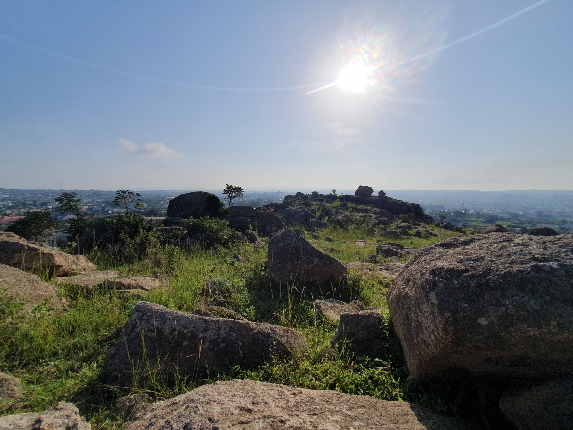 This is a wide-angle, eye-level shot of a rocky, overgrown hilltop overlooking a sprawling city. The sun is high in the sky, casting bright light and creating lens flare, suggesting it is midday. The foreground is dominated by large, weathered boulders interspersed with tall, green grass and some smaller shrubs. In the mid-ground, the terrain continues to be rocky with more vegetation, and a few sparse trees are visible. A thin, wispy cloud is present in the sky.
Beyond the hilltop, the view extends to a vast urban landscape. The city is densely populated with a mix of buildings, some appearing to be residential with visible rooftops. The distant horizon is hazy, typical of a large city. The overall impression is one of a natural outcrop within or on the edge of an urban environment. The specific location is the rocky summit of Gura Top in Nigeria, a prominent natural feature offering panoramic views of the surrounding city. No people or animals are visible in the scene.