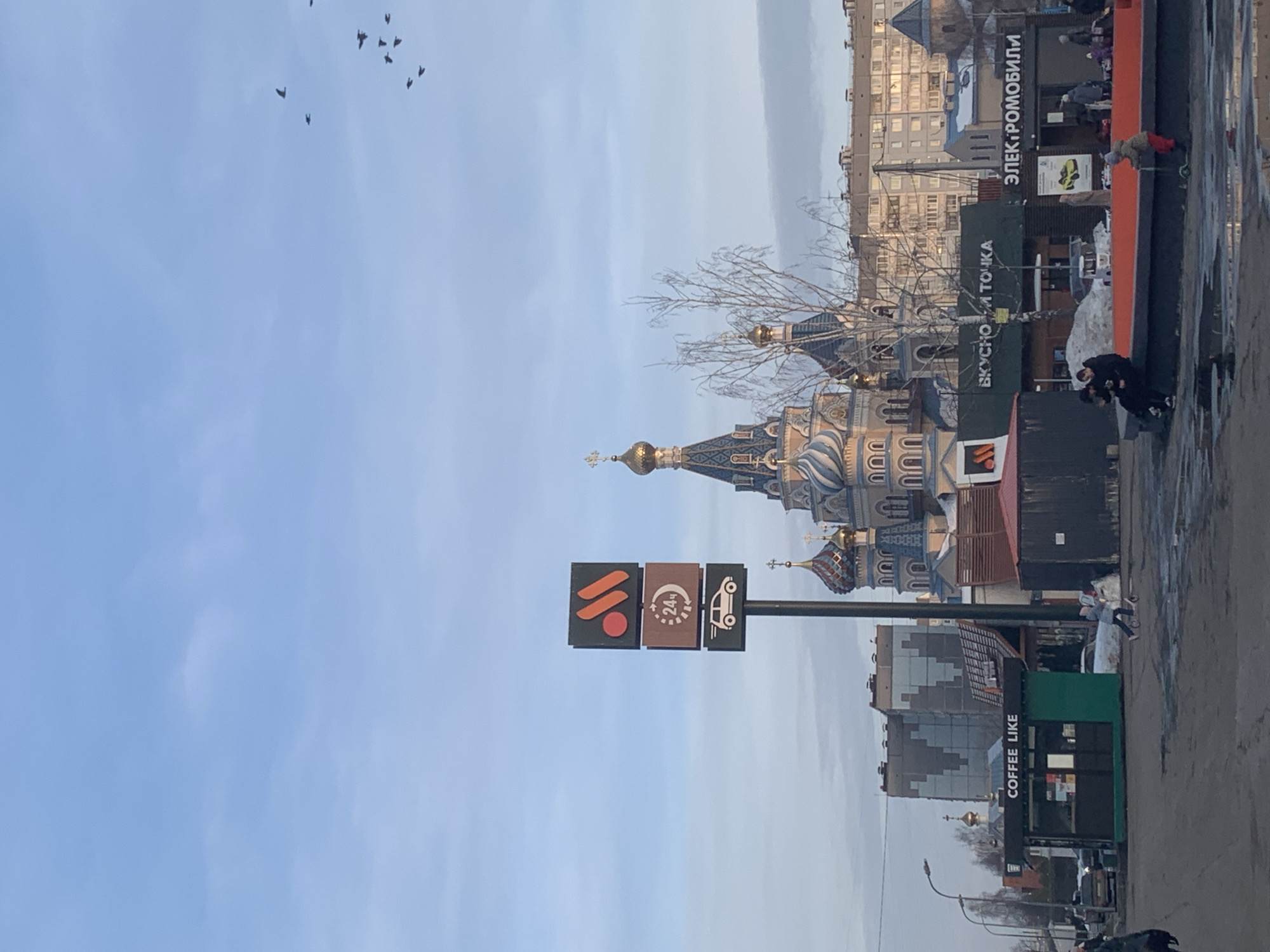 An outdoor, daytime urban scene in Izhevsk, Russia, presents a street-level view under a partly cloudy blue sky. In the mid-ground, the ornate St. Michael's Cathedral dominates, featuring multiple onion domes adorned in gold, blue, and patterned designs.
In the foreground, slightly to the left of center, a tall, freestanding sign for "Вкусно и точка" (Vkusno i tochka) displays its distinctive logo (red circle and orange stylized 'M' shapes), a "24ч" (24 hours) operation indicator, and a drive-thru car icon. Beneath this sign, various commercial kiosks are visible: a green and black "COFFEE LIKE" kiosk on the far left, another structure displaying "ВКУСНО И ТОЧКА" signage, and a kiosk further right marked "ЭЛЕКТРОМОБИЛИ" (Electric Cars).
The ground is a mix of wet asphalt or dirt, showing puddles and patches of melting snow, indicative of cold weather. A bare deciduous tree stands between the commercial kiosks and the cathedral.
Several individuals are present: In the lower right, two persons are seated on a low, red-orange concrete structure. A child in a light jacket and red pants is operating a green kick scooter on the wet ground near them. Other pedestrians are visible walking near the kiosks and buildings in the mid-ground. A flock of dark birds is in flight in the upper right portion of the sky.
Background elements include multi-story residential or commercial buildings, streetlights, and parked vehicles. The scene is illuminated by natural light, suggesting early morning or late afternoon.