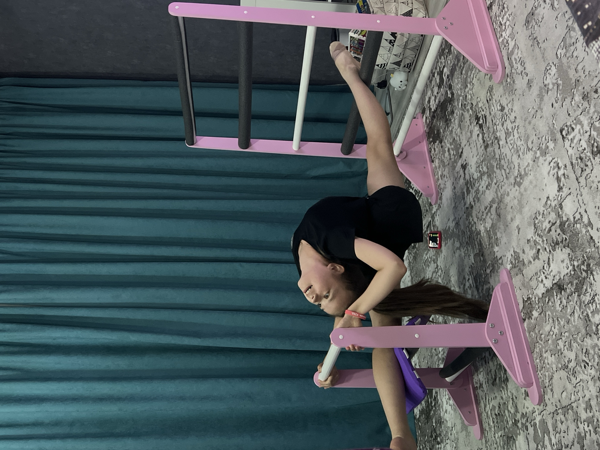 A young girl is doing a split on a pink and white indoor gymnastic ladder. She is wearing black shorts and a black t-shirt. She is holding onto a ladder rung with her hands and has one leg on the upper rungs of the ladder. The setting appears to be indoors, near a teal-colored curtain and a patterned gray and white floor. The exact location is not discernible from the image. It is not possible to ascertain the city of Chaikovsky, Russia, based solely on the visual information presented.