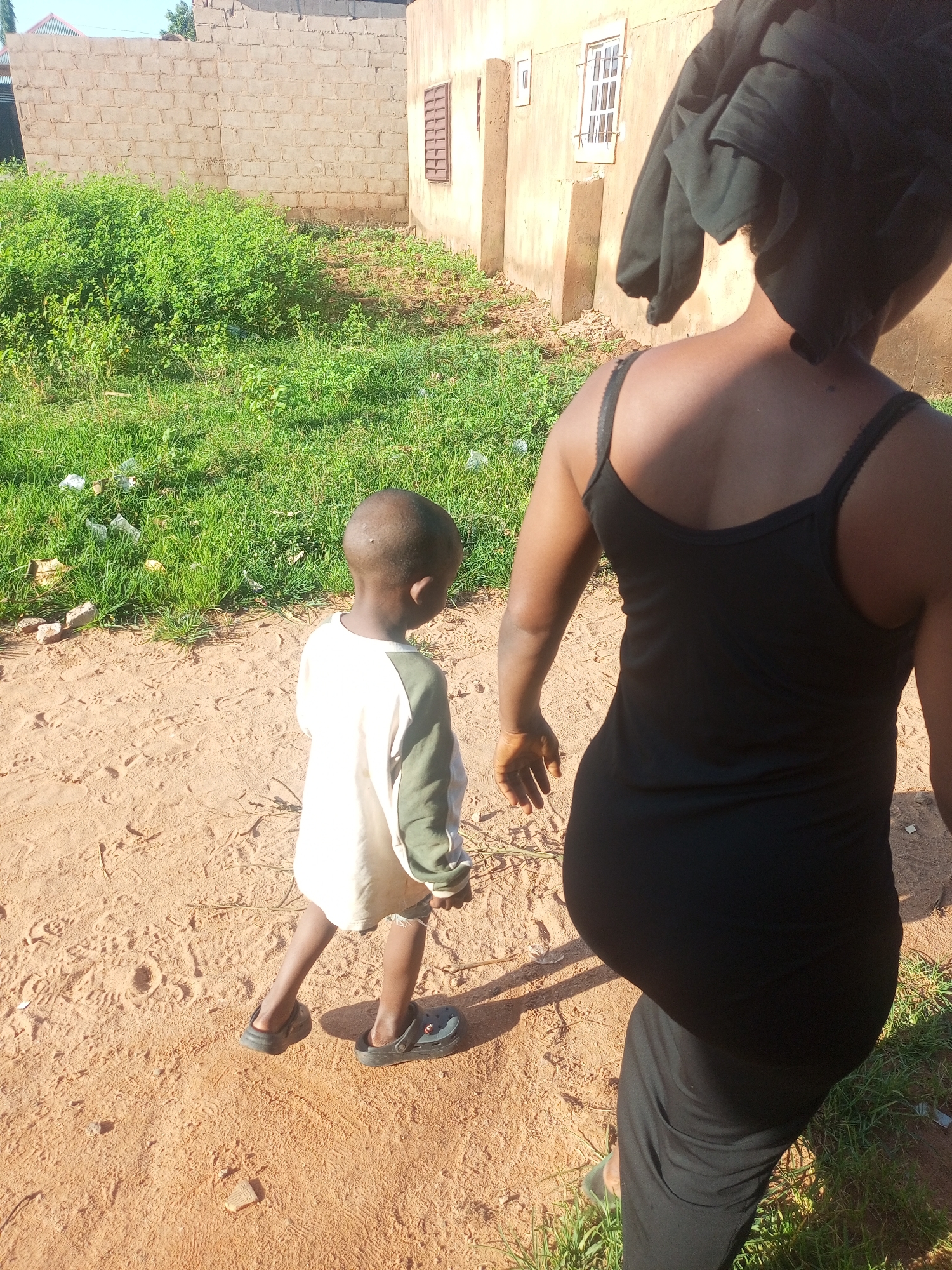 Two individuals, an adult and a child, are walking away from the camera on a sandy path. The adult, positioned closer to the camera and on the right side of the frame, is wearing a black sleeveless dress and a black headwrap. The child, on the left, is wearing a long-sleeved shirt with a white front and green sleeves, along with dark colored clogs. Behind them, a building with a light brown facade and a barred window is visible. The ground is a mixture of sand and patches of green grass, with some scattered debris. The overall environment appears to be an outdoor urban setting in Tempure, Nigeria.