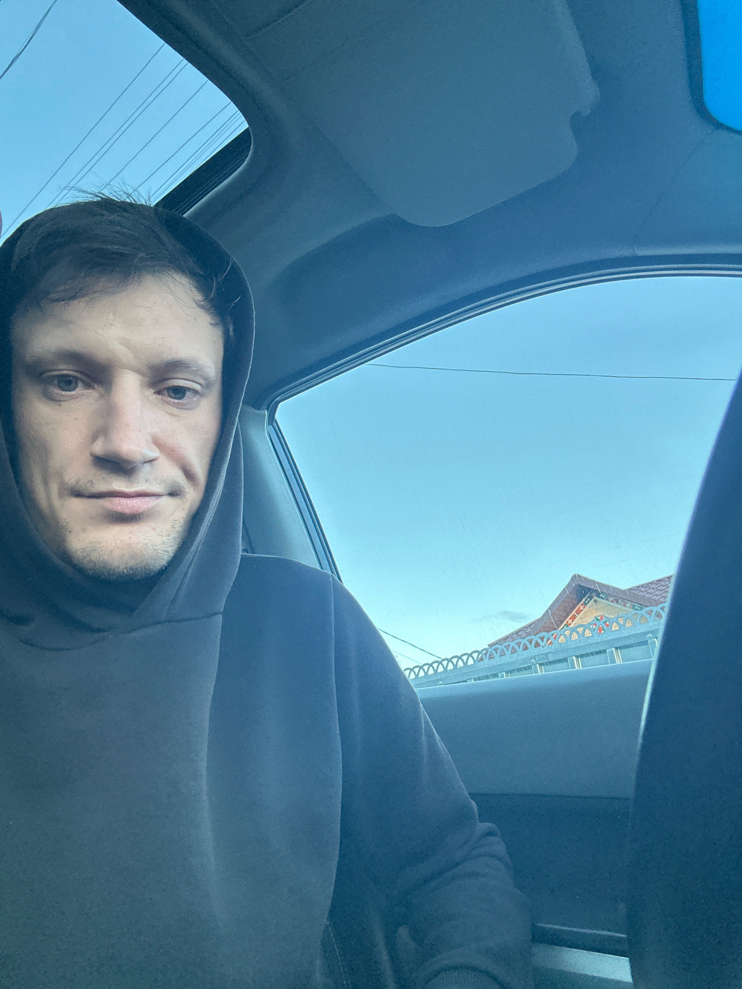 The image is a selfie taken from inside a car, looking towards the front passenger seat. A man with short, dark hair and light-colored eyes is wearing a dark hoodie and looking directly at the camera. The car's sunroof is open, revealing a clear blue sky with some power lines visible. Through the passenger side window, a building with a tiled roof and decorative fence is visible, suggesting an urban or suburban setting. The man appears to be in Makiv, Ukraine, based on the provided context.