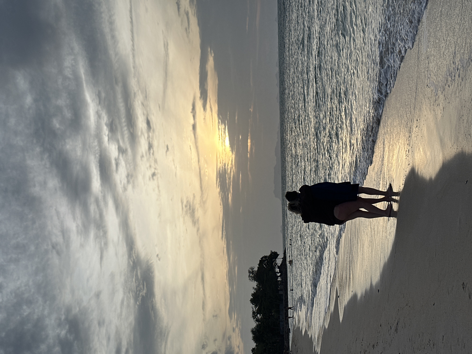 Two individuals, silhouetted against a dusky sky, embrace on a sandy beach in Ko Samui District, Thailand. The person on the left, wearing a dark top and patterned bottoms, has their left arm around the shoulders of the person on the right, who is wearing a dark top and dark bottoms. They stand facing the ocean, their feet submerged slightly in the shallow wash of waves. The sun, partially obscured by dramatic, streaky clouds, casts a warm, golden light across the water and the sand, creating a long shadow from the couple. The ocean is calm, with gentle waves lapping the shore. In the background, a line of dark green trees forms a silhouetted treeline, with a few indistinct figures visible on the beach further down. Distant islands or landmasses are faintly visible on the horizon. The overall atmosphere is serene and romantic, with the natural beauty of the sunset and the ocean setting a tranquil mood.