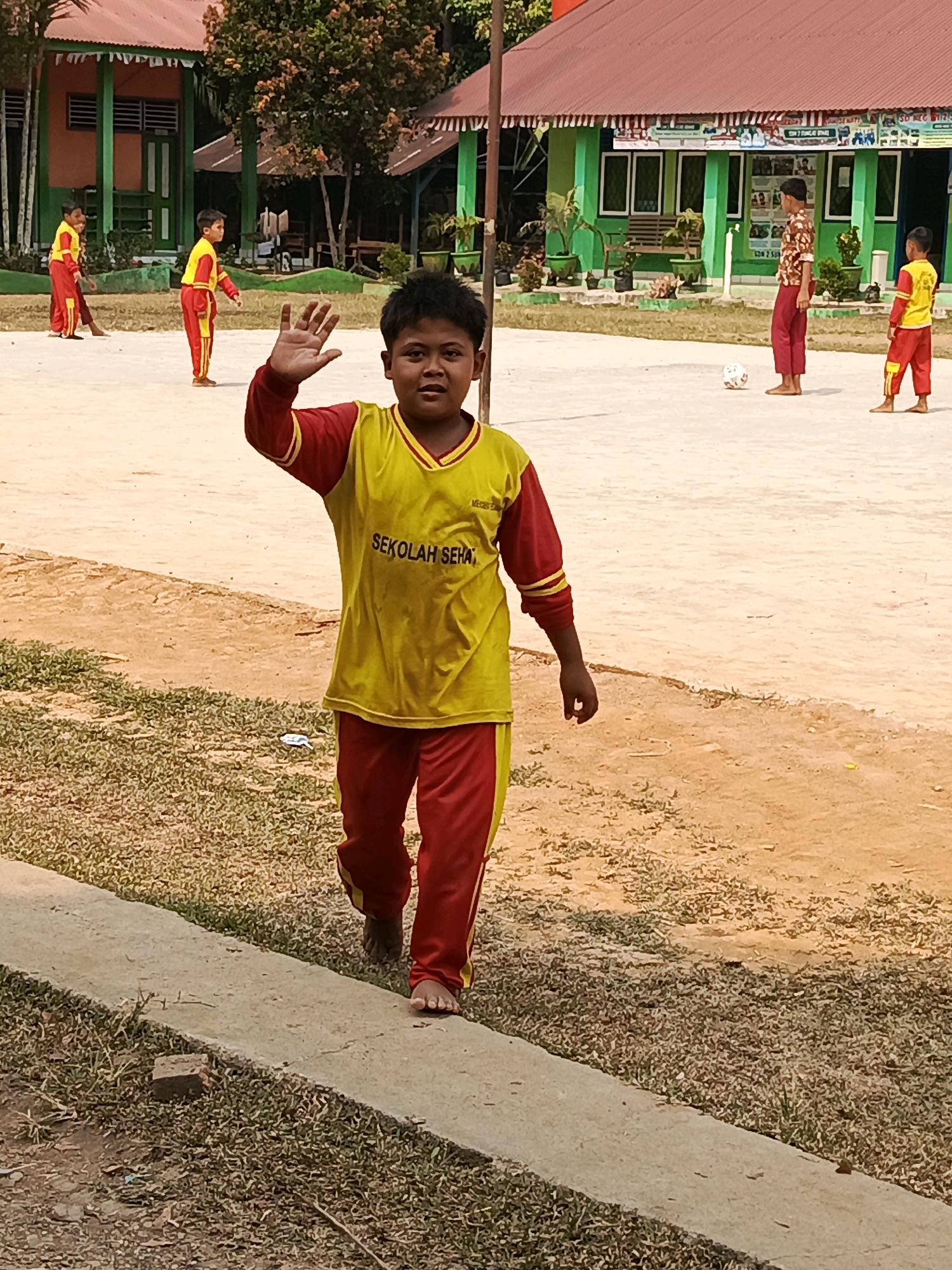 The image depicts a sunny outdoor scene at a school in Megang Sakti IV, Indonesia. In the foreground, a young boy wearing a yellow and red school uniform with the text "SEKOLAH SEHA" on his shirt is waving with his right hand. He is looking towards the camera and appears to be walking. His uniform consists of a yellow t-shirt with red sleeves and collar, and red pants with yellow stripes. His left hand is resting at his side.
In the middle ground, several other children in similar red and yellow uniforms are present. Two children are standing further back on the left, while another two are on the right. One of the children on the right is interacting with a soccer ball. Behind the children, a green building with a red roof is visible, which appears to be part of the school. The ground is a mix of sandy dirt and grass. The overall atmosphere is that of a schoolyard during an activity.