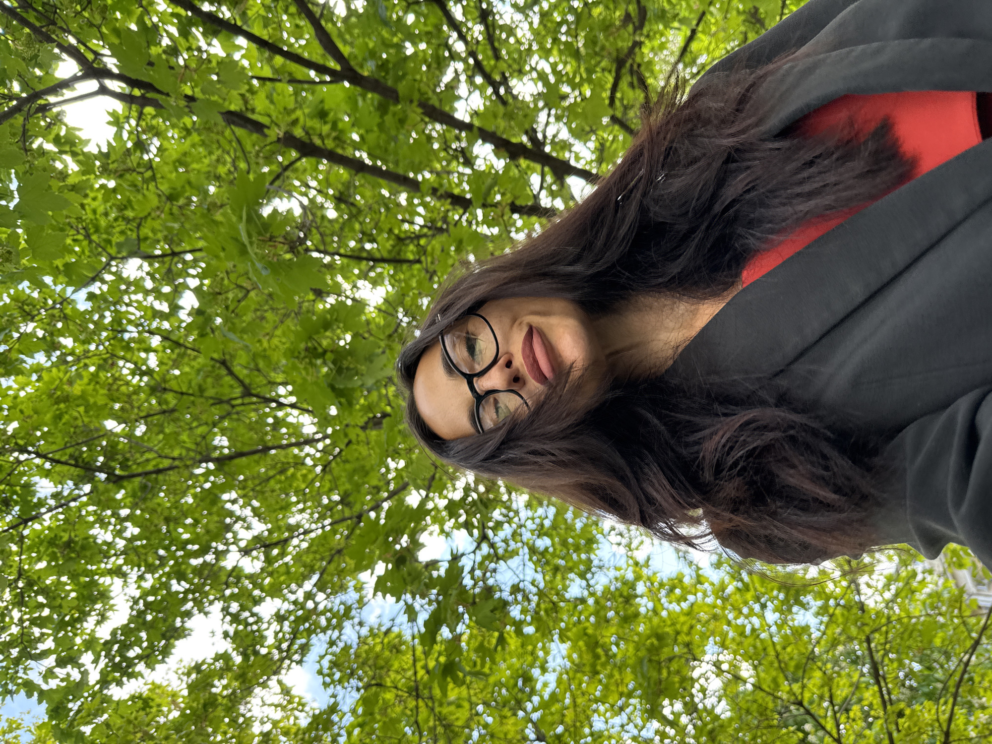 A low-angle, outdoor shot of a woman wearing eyeglasses and a dark jacket over a red top. Her long, dark hair is partially obscuring her face, and she has her tongue slightly out. The background is filled with green foliage and branches, suggestive of trees overhead. The scene is possibly set in the city of Pleven, Bulgaria, though specific location details are not discernible from the image itself.
