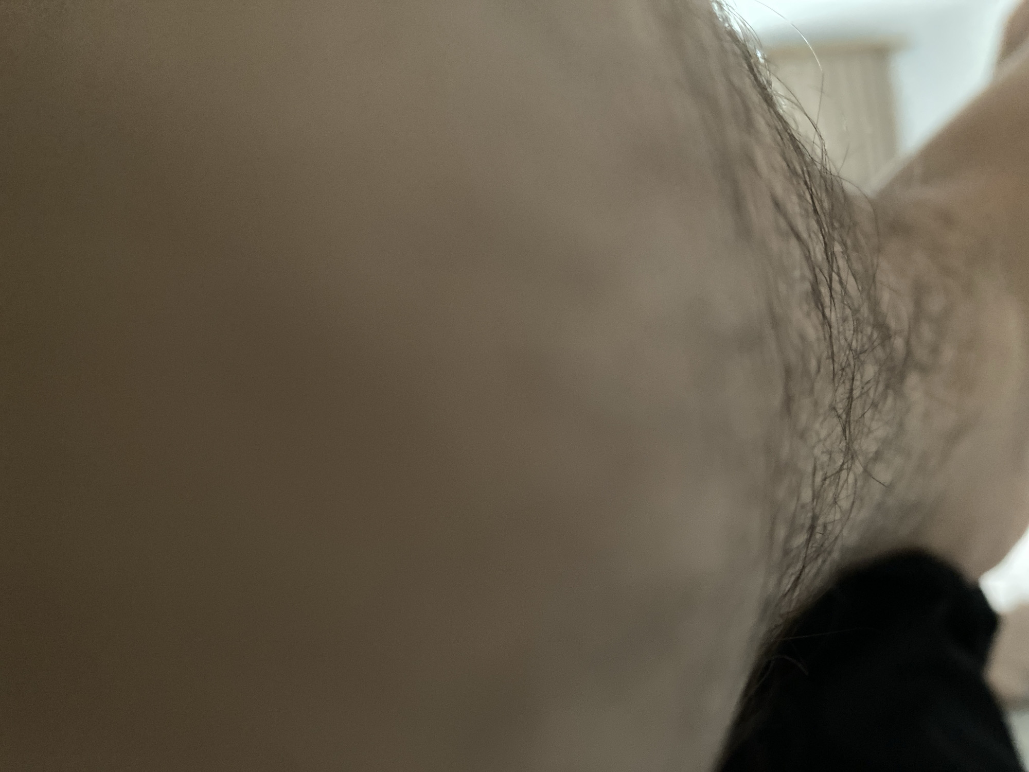 The image is a close-up, distorted, and abstract view of a person's armpit hair, possibly taken from a very low angle. The lighting is dim and uneven, with some areas appearing brighter than others. The background is blurred and indistinct, with hints of what might be indoor lighting and possibly furniture. There are no discernible features that would allow for the identification of a specific location within the city of Qaraoy, Kazakhstan. The focus is on the texture and fine details of the hair and skin.