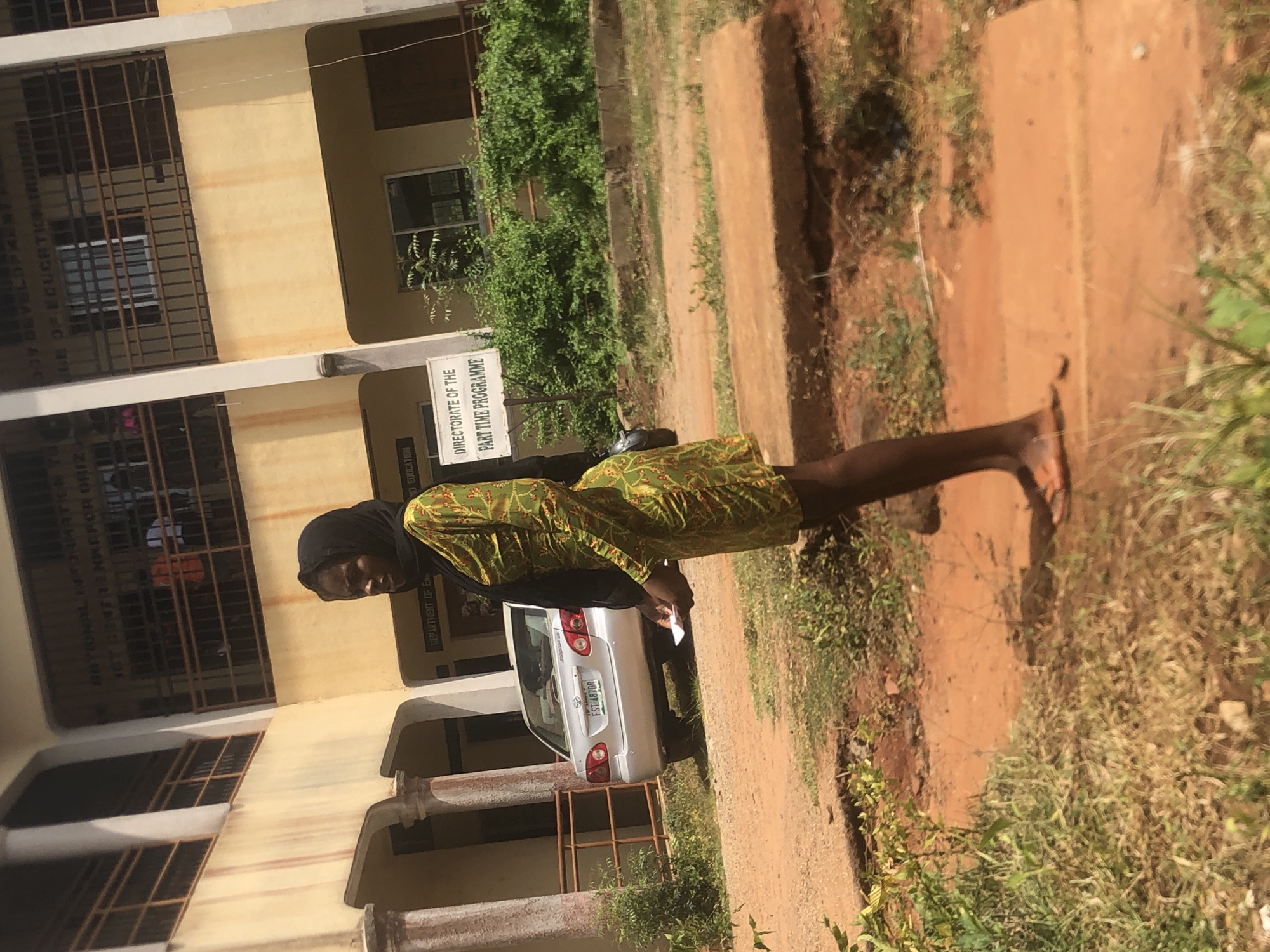 The image shows a woman in a headscarf and patterned dress standing next to a silver Toyota Corolla with the license plate FST-487DR. The woman appears to be holding a piece of paper. The car is parked in front of a building which houses the "Directorate of the Part Time Programme", and the “Department of Education”. The photo may be near or at the University of Port Harcourt Open and Distance Learning Centres in Ukwu-Abwa, Nigeria, based on the information on the signs and the license plate. The surroundings consist of greenery and what appears to be a dirt or gravel path. The building appears to be multi-storied with barred windows. The lighting indicates it is daytime.