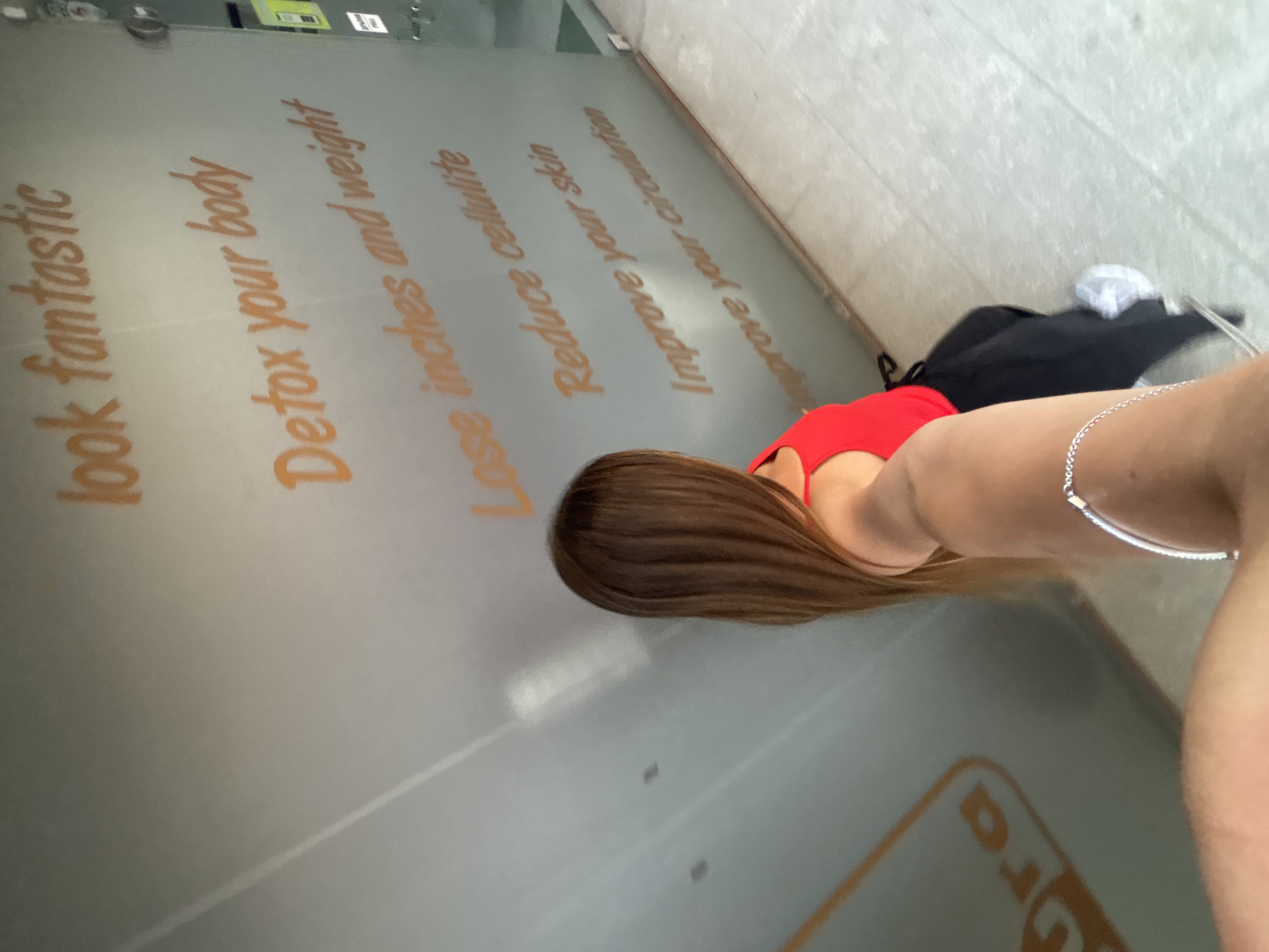 A woman in a red top and black pants is standing near a wall with orange lettering in Sofia, Bulgaria. The text on the wall includes advertising copy related to wellness: "look fantastic", "Detox your body", "Lose inches and weight", "Reduce cellulite", "Improve your skin", and "Improve your circulation". A black bag and white shoes are on the ground. The lower part of a commercial logo is partially visible on the wall.