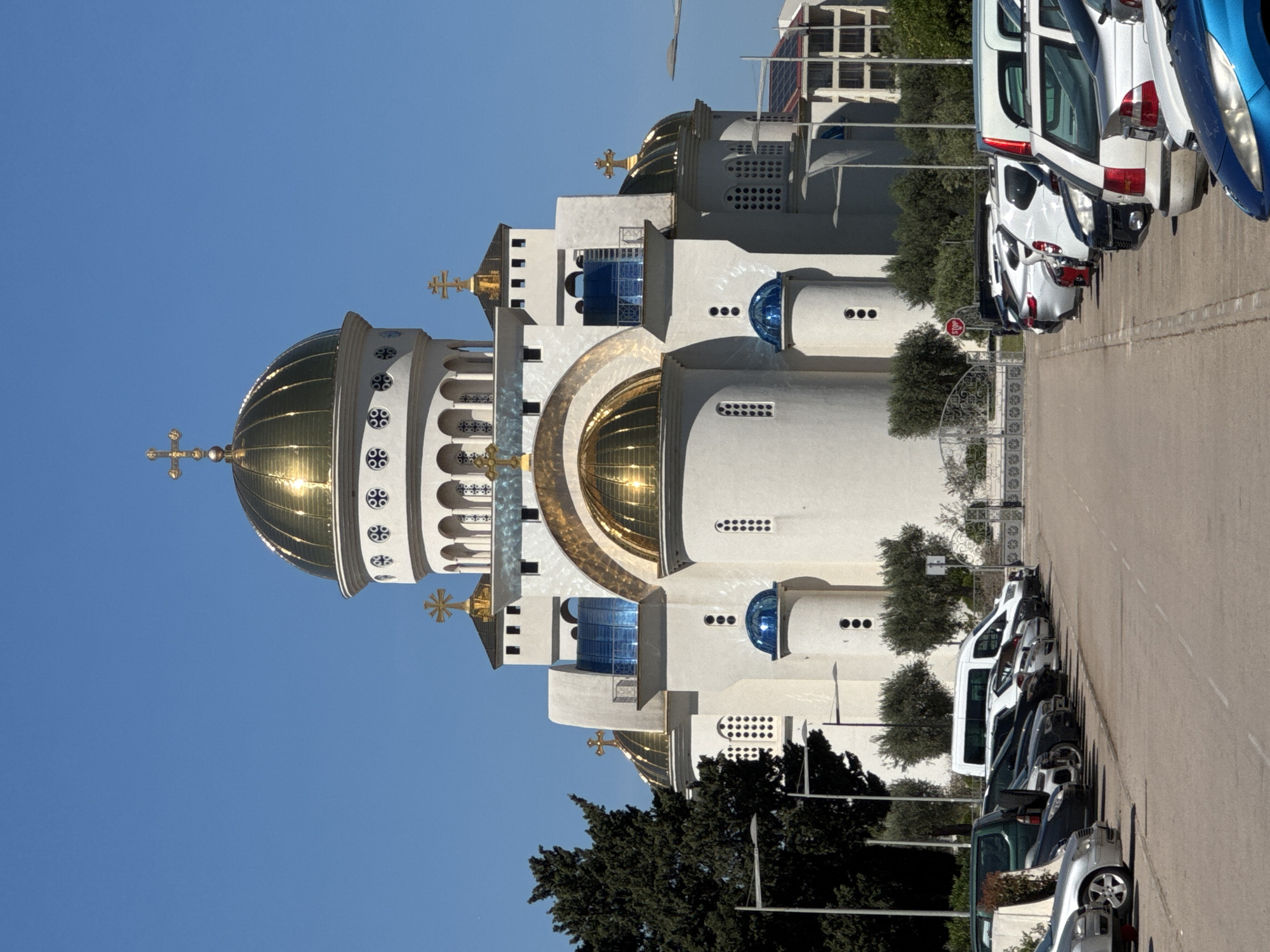 The image shows the Saint John Vladimir Orthodox Cathedral in Bar, Montenegro. The cathedral is white with gold domes. Parked vehicles are along the right side of the building. The sky is blue. Some trees and shrubbery are near the building. A stop sign is visible.