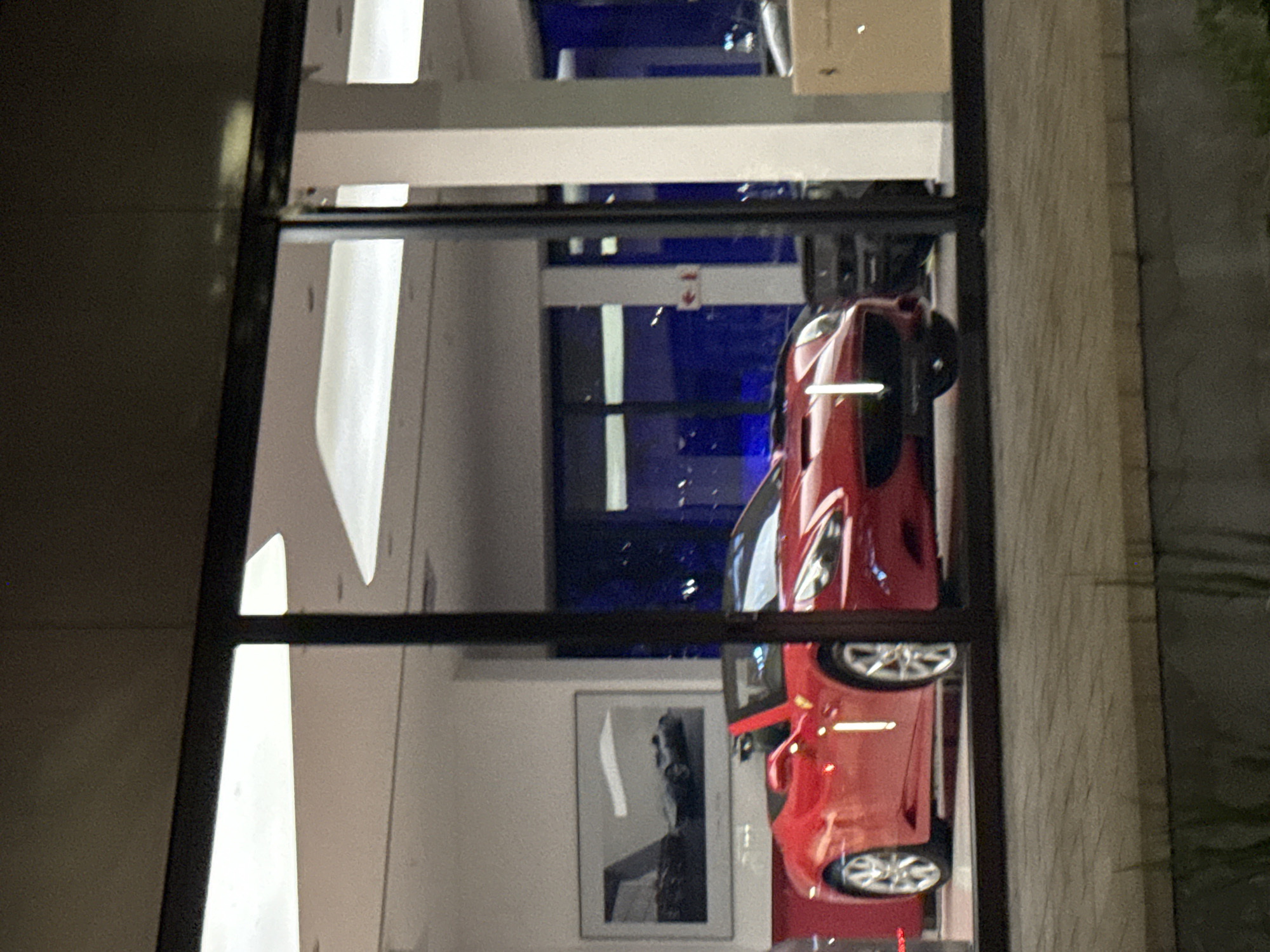 The image shows a red sports car behind a window. The car is the main subject, with details like its wheels and sleek design visible. The window creates a reflective surface, revealing elements of the interior, including a framed picture. The location is suggested to be Cape Town, South Africa, although no explicit landmarks confirm this. The environment is urban, indicated by the building and surrounding structures. The scene appears to be a car dealership or showroom, showcasing the vehicle.