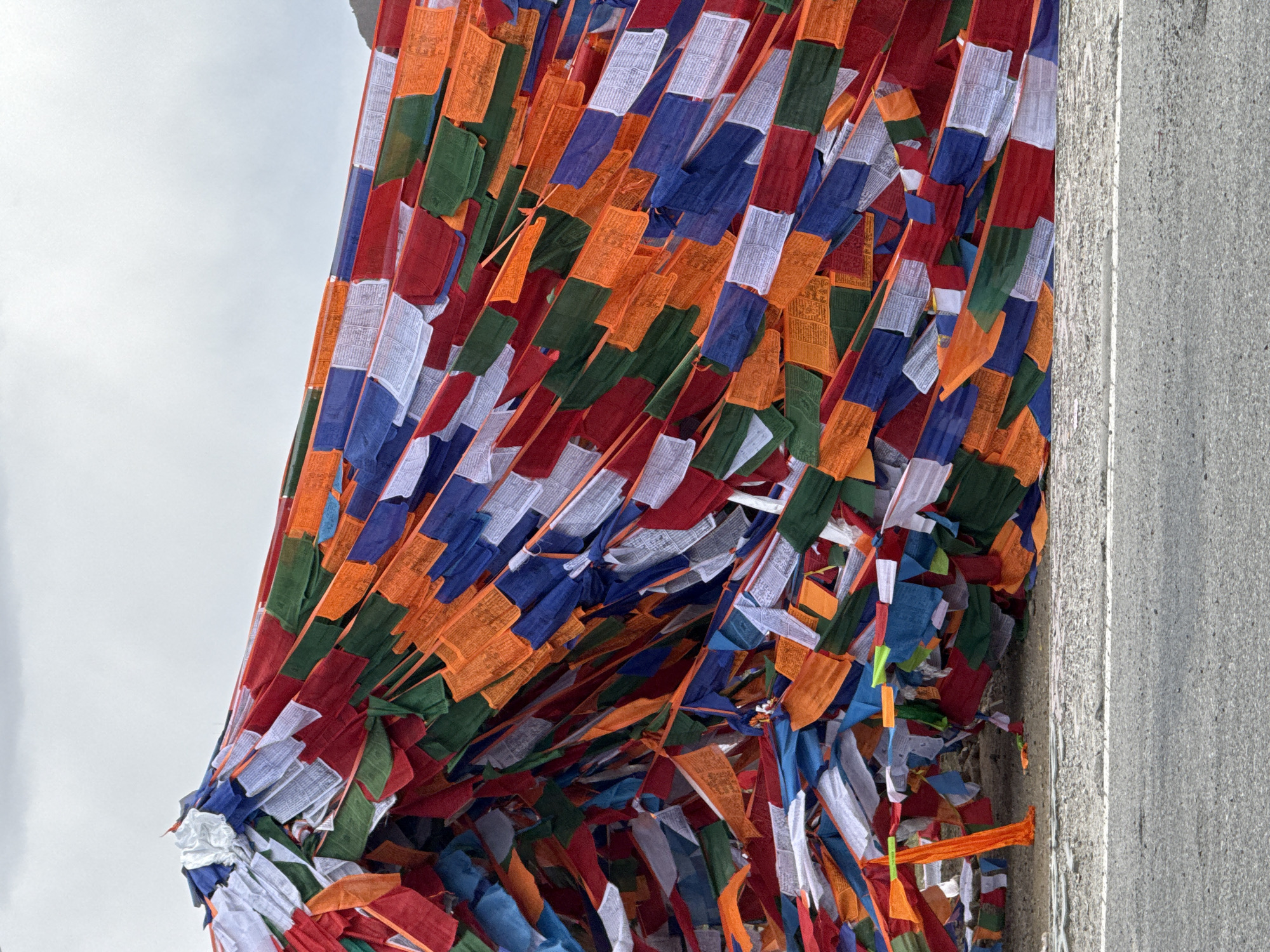 A vibrant and dense array of Buddhist prayer flags dominates the image, stretching across the frame against a backdrop of a light sky and a textured grey wall. The flags are strung horizontally in multiple layers, creating a visually rich tapestry of color and texture. They are predominantly in the five traditional colors: blue, white, red, green, and orange (often representing yellow or earth).
Each flag is a small, rectangular piece of fabric, and many display intricate black or dark-colored text and symbols, likely prayers or mantras, which are partially visible upon closer inspection. The flags appear to be fluttering, suggesting the presence of a gentle breeze or wind.
To the left of the frame, a bright, overcast sky provides a light background for the colorful flags, indicating that the photo was taken during the daytime. On the right side, a solid, grey-toned wall or structure, possibly concrete, runs vertically, acting as an anchor or boundary for the strung flags. The overall impression is one of spiritual offering and cultural tradition, characteristic of regions influenced by Tibetan Buddhism, such as Lhatsé, China.