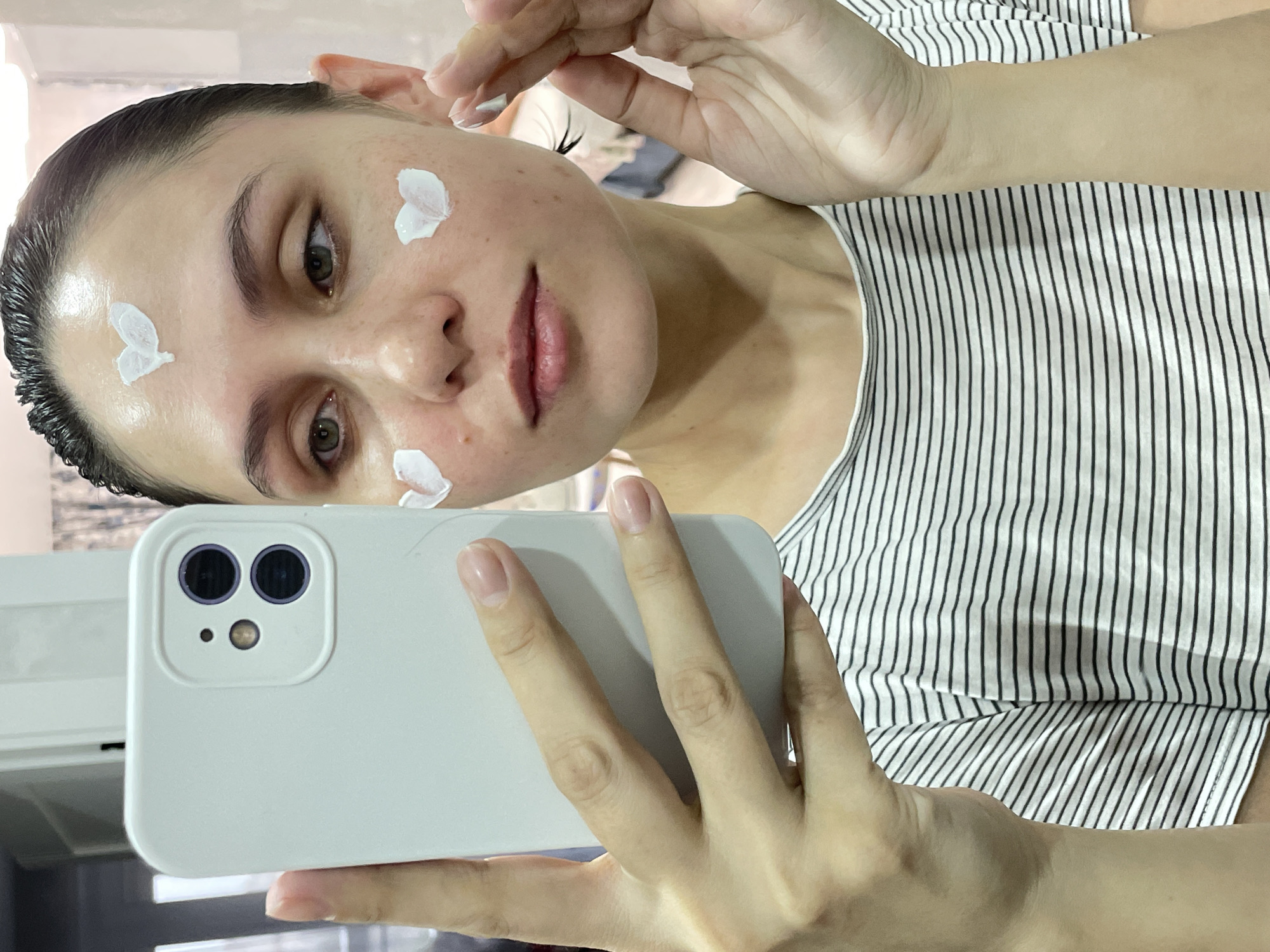 The image shows a person with fair skin and dark hair, taking a selfie. They are wearing a striped black and white shirt and have dabs of white cream on their forehead, cheek, and face. The person is holding a white smartphone in their left hand, with the camera lenses visible. The background is blurry but appears to be an indoor setting. There is no information in the image to indicate this photo was taken in Izhevsk, Russia.