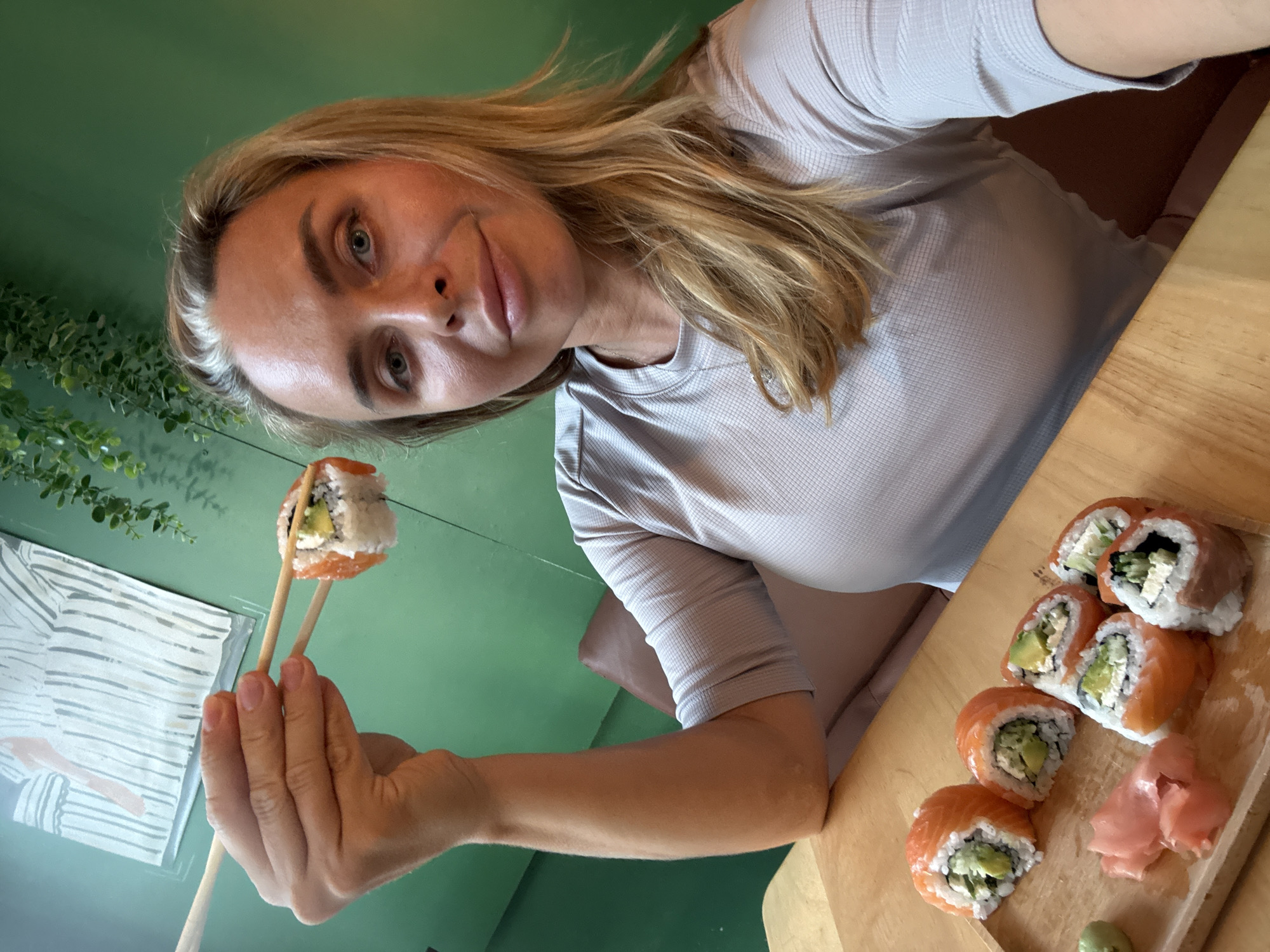 A blonde woman is holding a piece of sushi with chopsticks. She is sitting at a wooden table in a restaurant. There are more pieces of sushi on the table, along with some pickled ginger and wasabi. The restaurant has green walls and a piece of art on the wall. The woman is wearing a grey shirt. The scene is indoors and appears to be in Baan Bo Phut, Thailand.