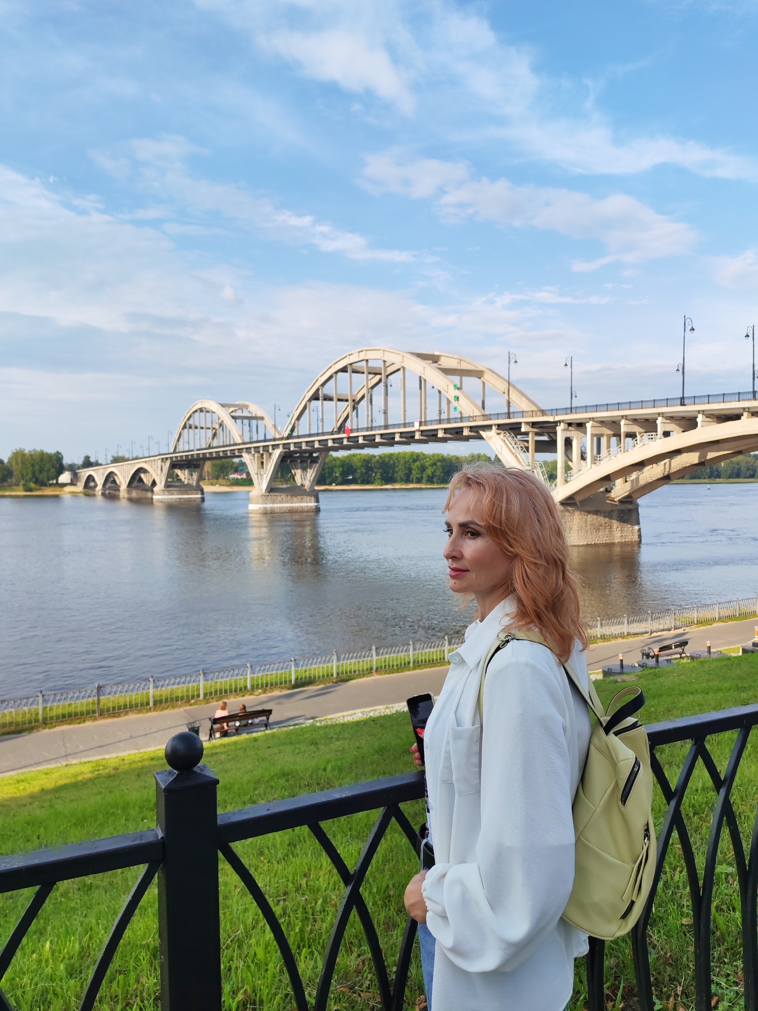 A woman with reddish-blonde hair stands in the foreground, facing right, with a city bridge and a wide river in the background. She wears a white oversized shirt and a light yellow backpack over her right shoulder. In her left hand, she holds a dark smartphone. Behind her, a black metal fence borders a grassy slope leading down to a paved walkway along the river. On the walkway, two people are seated on a bench, and several lampposts are visible. The bridge, a large concrete structure with arched spans, crosses the river under a partly cloudy blue sky. The setting appears to be an urban park or promenade in Bol'shie Mkhi, Russia.