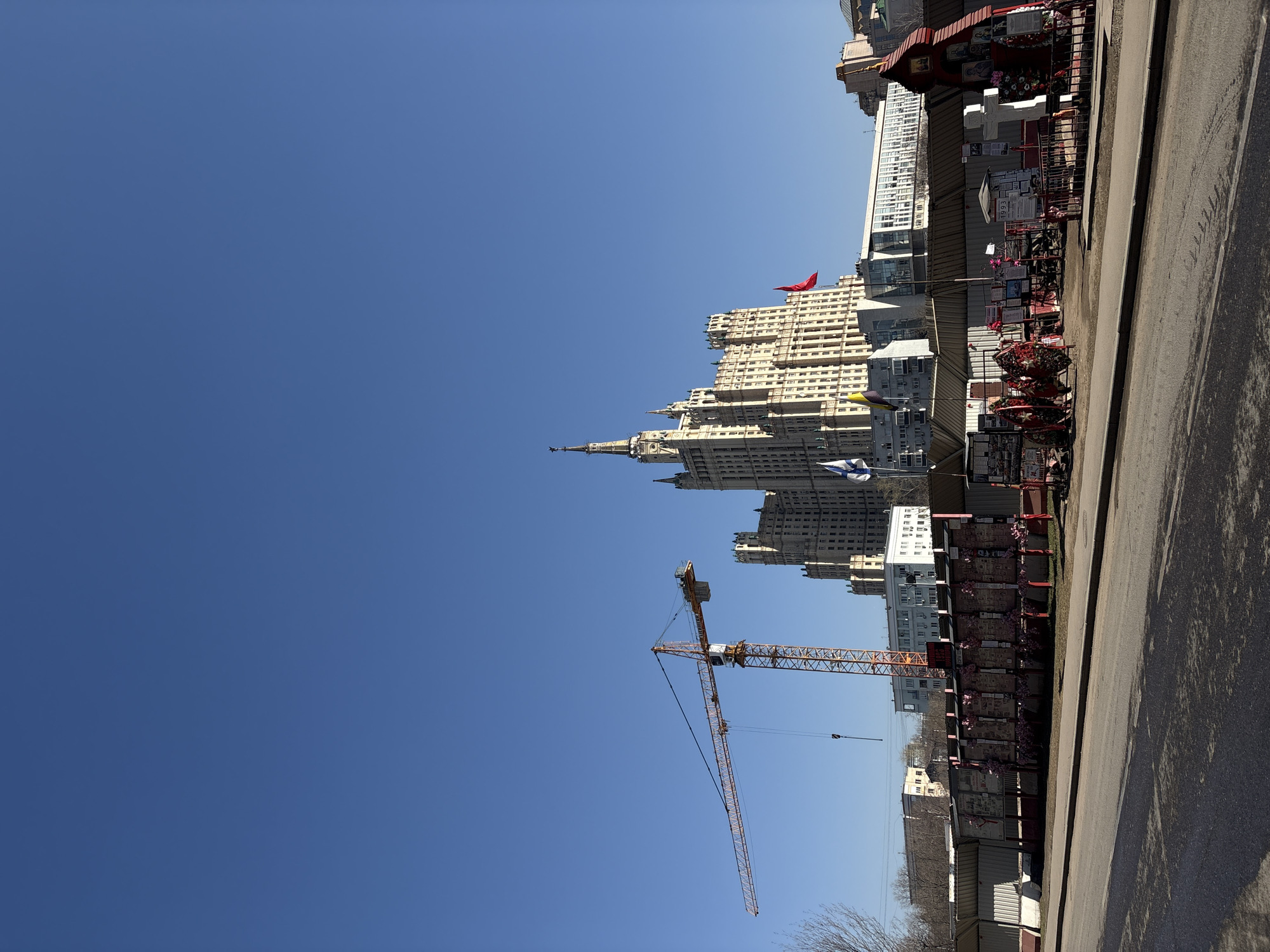 A vertical, medium-shot photograph captures a sunny urban scene in Moscow, Russia, featuring a mix of architectural landmarks and construction elements against a clear blue sky. In the foreground and stretching along the right side of the frame is an asphalt road. Adjacent to the road is a collection of memorials and structures, possibly makeshift shrines or displays.
To the left of the road and directly ahead stands the Ministry of Foreign Affairs building, one of the "Seven Sisters," a series of Stalinist skyscrapers. The building's distinctive architectural features, including its spired tower and ornate facade, are clearly visible.
A construction crane is positioned to the left and slightly in front of the Ministry of Foreign Affairs building. The crane is red-orange and has a lattice structure. In the lower foreground, some trees are visible, suggesting a park or green space. The image suggests a city undergoing continuous change, where historical landmarks exist alongside modern construction.