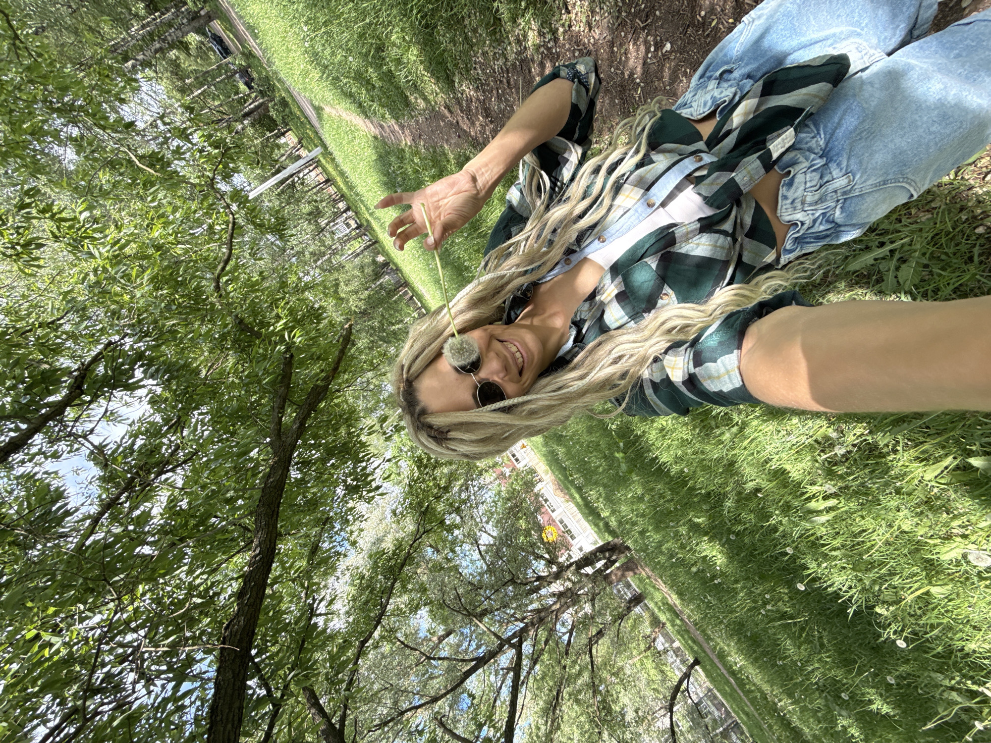 A woman lies in the grass in Rusanovo, Russia, holding a dandelion. She wears light-wash jeans, and a green and black plaid shirt. The park is visible in the background, with trees and greenery.