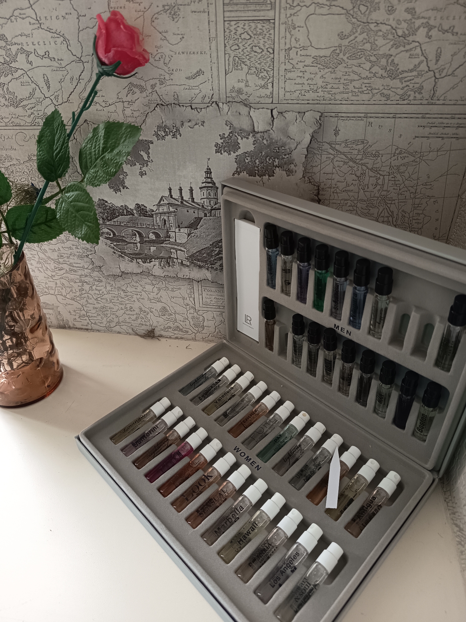 The image shows a collection of perfume samples neatly arranged in a grey case. The case is divided into sections, with some labeled "MEN" and "WOMEN". The perfume bottles are small and clear, with white caps. Each bottle has a label with a name, such as "Santorini", "Romance", "Marbella", "Hawaii", "Los Angeles", and "Boston". Some labels also indicate "EAU DE PARFUM" or "COLLECTION".
In the background, a vintage map is visible, which appears to be a decorative wallpaper. A single artificial red rose with green leaves in a copper vase stands to the left of the perfume case. The setting appears to be indoors, likely on a table or shelf, with natural light casting shadows. The overall mood is one of curated presentation, possibly a gift set or a personal collection of fragrances. The text present is primarily on the perfume labels and the background map.
