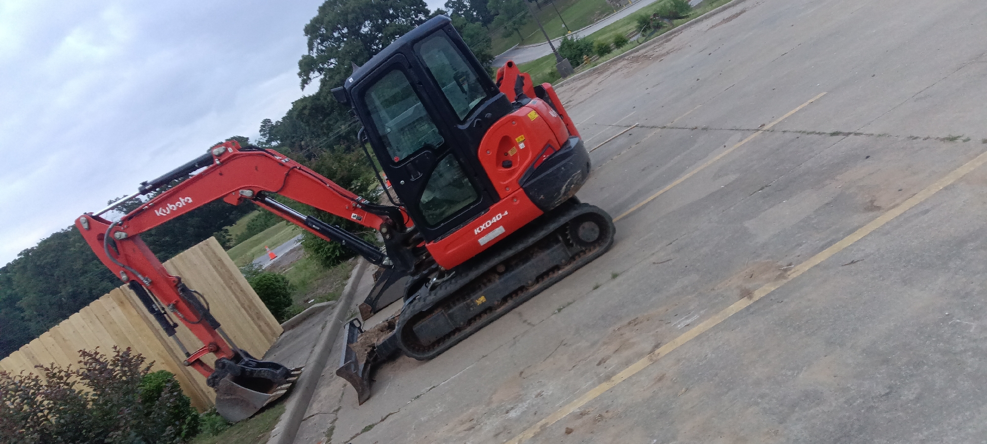 The image depicts a red and black Kubota KX040-4 mini excavator positioned on an asphalt surface marked with yellow parking lines. The excavator's boom is extended to the left, with the bucket near a wooden fence and some shrubbery. The background includes trees and what appears to be a commercial or public building, possibly in the city of Van Buren, Arkansas, USA. The sky is overcast. The excavator itself appears stationary.