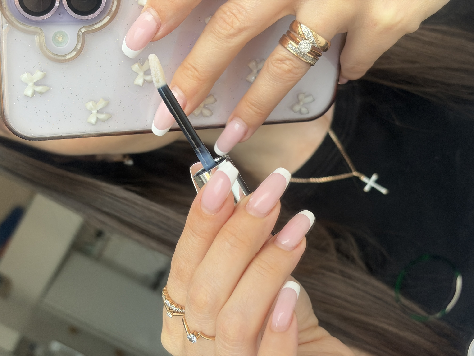 A close-up shot shows a person holding a makeup applicator, possibly for lip gloss or liquid lipstick, in their right hand with perfectly manicured nails with a classic French manicure, consisting of a pale pink base and a bright white tip. The person's hand is also adorned with a gold ring with several rows of diamonds. The hand is holding a white glitter phone case. Another hand with similar manicured nails and gold jewelry can be seen as well. Part of the person's shoulder, hair, and necklace with a cross pendant are visible. The photo does not include location specifics related to the city of Saint Petersburg, Russia.