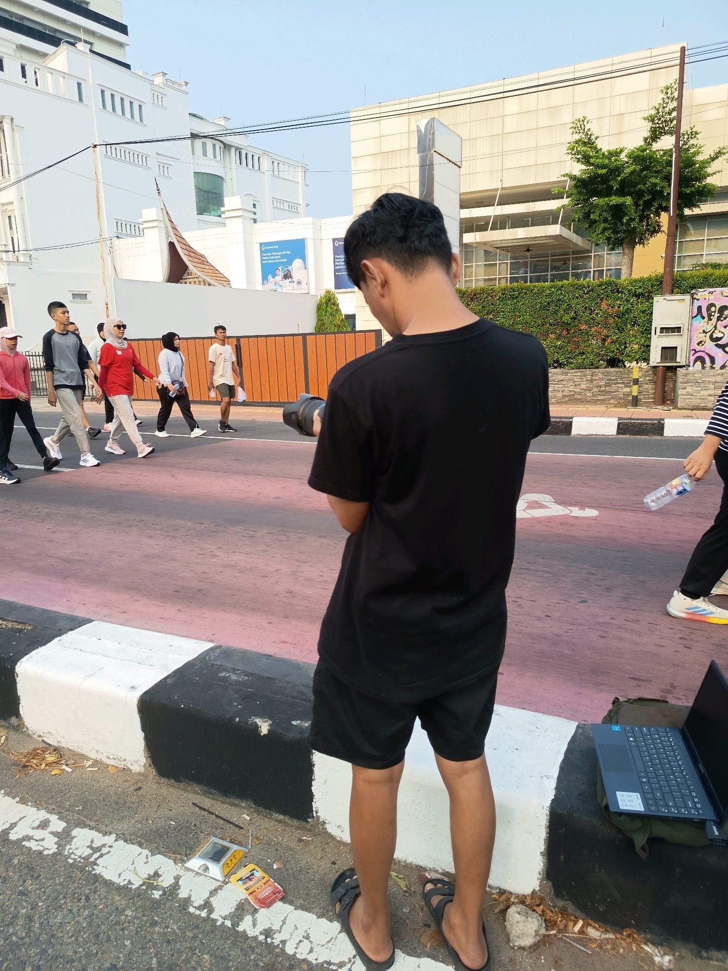 In Padang, Indonesia, a young man is captured from behind, facing away from the camera and holding a professional camera with a lens. He is wearing a black t-shirt and black shorts, and casual sandals. To his left, several people are walking across a pink and white striped crosswalk. Among them are individuals wearing activewear, including a woman in a red shirt and hijab, and another woman in a grey long-sleeved shirt and black pants. A laptop sits open on a backpack on the ground to the right of the young man. The background features modern buildings, including one with a distinctive curved roofline and another with a large glass facade. Lush green foliage is also visible. The scene appears to be a public space in the city during daylight hours.