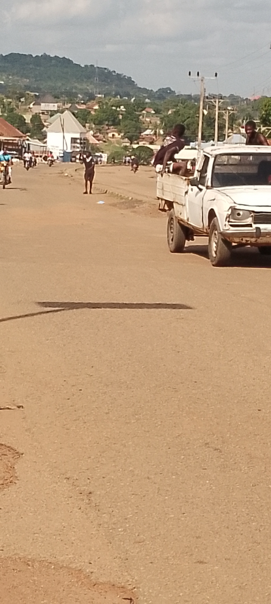In Gboko, Nigeria, a white pickup truck drives down a dusty, paved road. Several young men are riding in the bed of the truck, some leaning over the sides. One person stands in the middle of the road with their arms raised, looking towards the truck. In the background, buildings and trees are visible, with a large, green hill in the distance. The sky is overcast with clouds. The scene appears to be during the daytime.