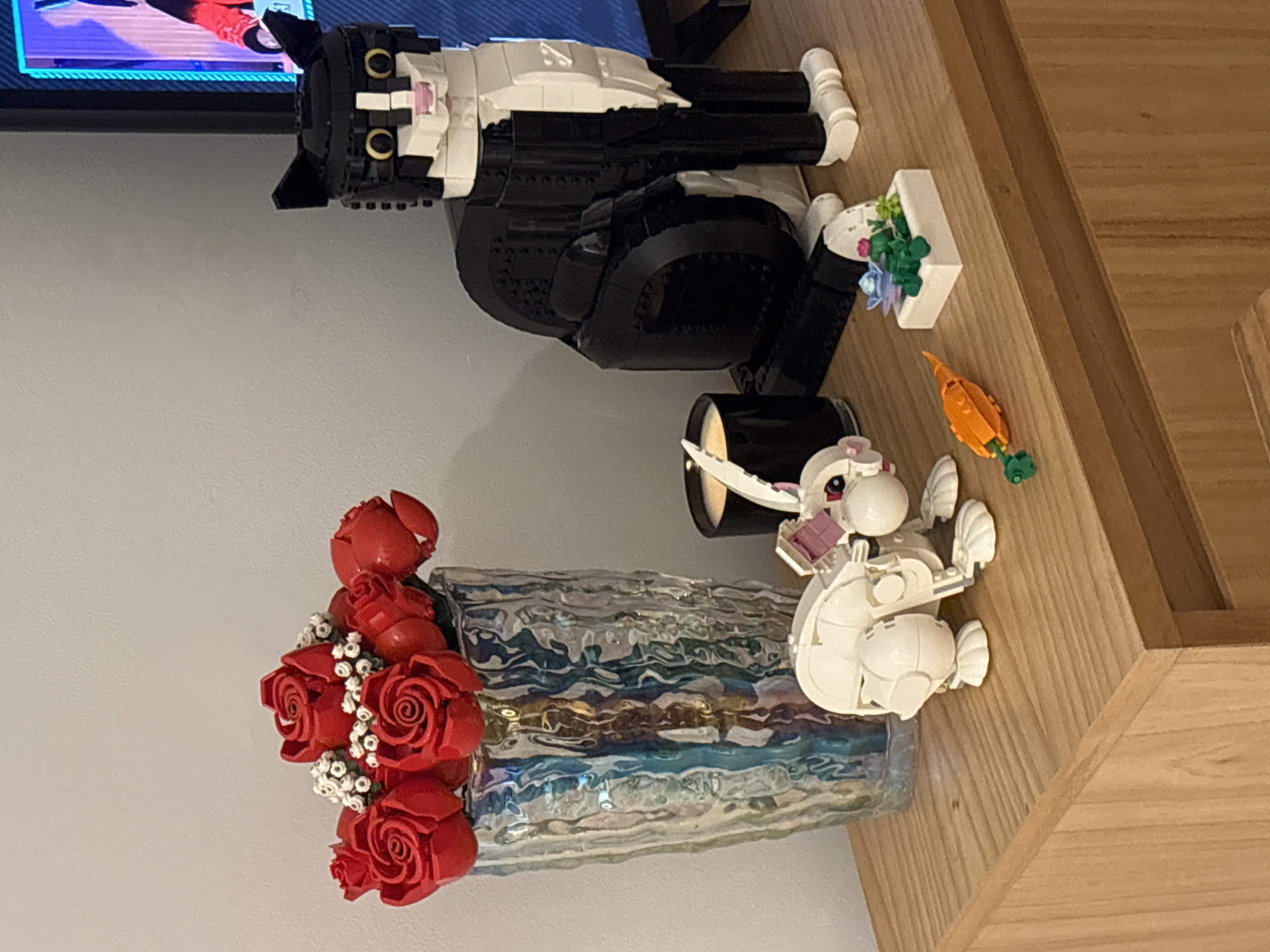 The scene presents a collection of objects arranged on a wooden surface. A vase filled with red roses sits on the left, made of interlocking plastic pieces. Above it, a constructed black and white cat figure made of interlocking plastic pieces reclines near a miniature constructed planter with green and blue elements. An orange interlocking plastic carrot and a white interlocking plastic rabbit are placed to the right of the vase. The background consists of a light gray wall and a television screen showing a colorful image. There is no evident location in Buenos Aires, Argentina.