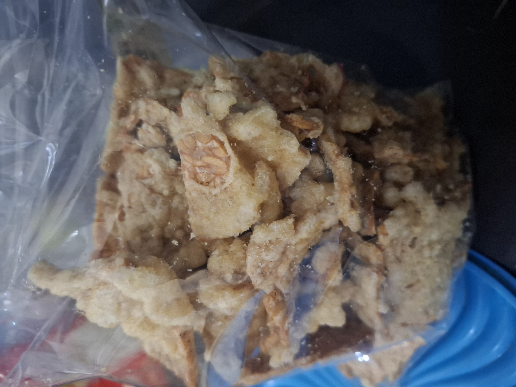 A close-up shot shows a clear plastic bag filled with what appears to be fried crispy snacks. The snacks are irregular in shape, light golden brown in color, and some have visible textures suggesting they might be made from shrimp or seafood. The bag is partially open at the top, revealing the contents. The background is dark, with a bright blue, ridged surface visible at the bottom left, possibly a plate or tray. The lighting is diffused, creating a slightly hazy appearance of the snacks through the plastic. There are no people or specific locations in the city of Maleber, Indonesia, identifiable in this image.