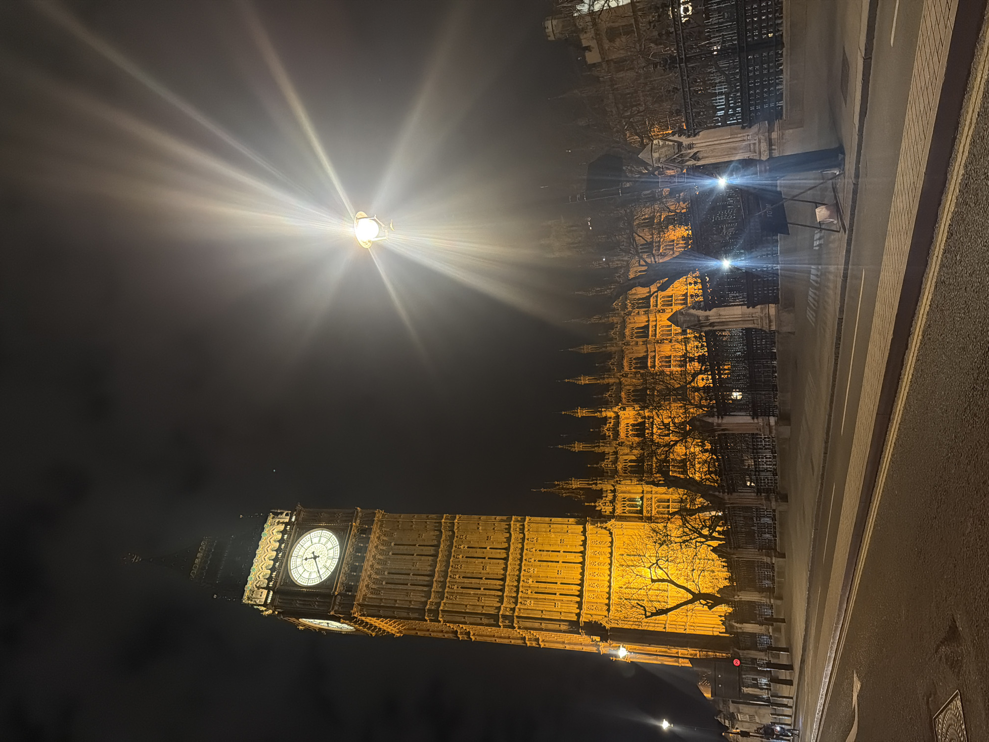 This photograph captures a night scene in London, United Kingdom, featuring the illuminated Elizabeth Tower (housing Big Ben) and the Houses of Parliament. The tower's clock face is visible. A street lamp shines brightly above, creating lens flare in the image. The buildings are lit up in golden light. A section of a street and a pavement are visible in the foreground. A security barrier is on the right side of the image along the street. Tree branches are visible between the buildings.