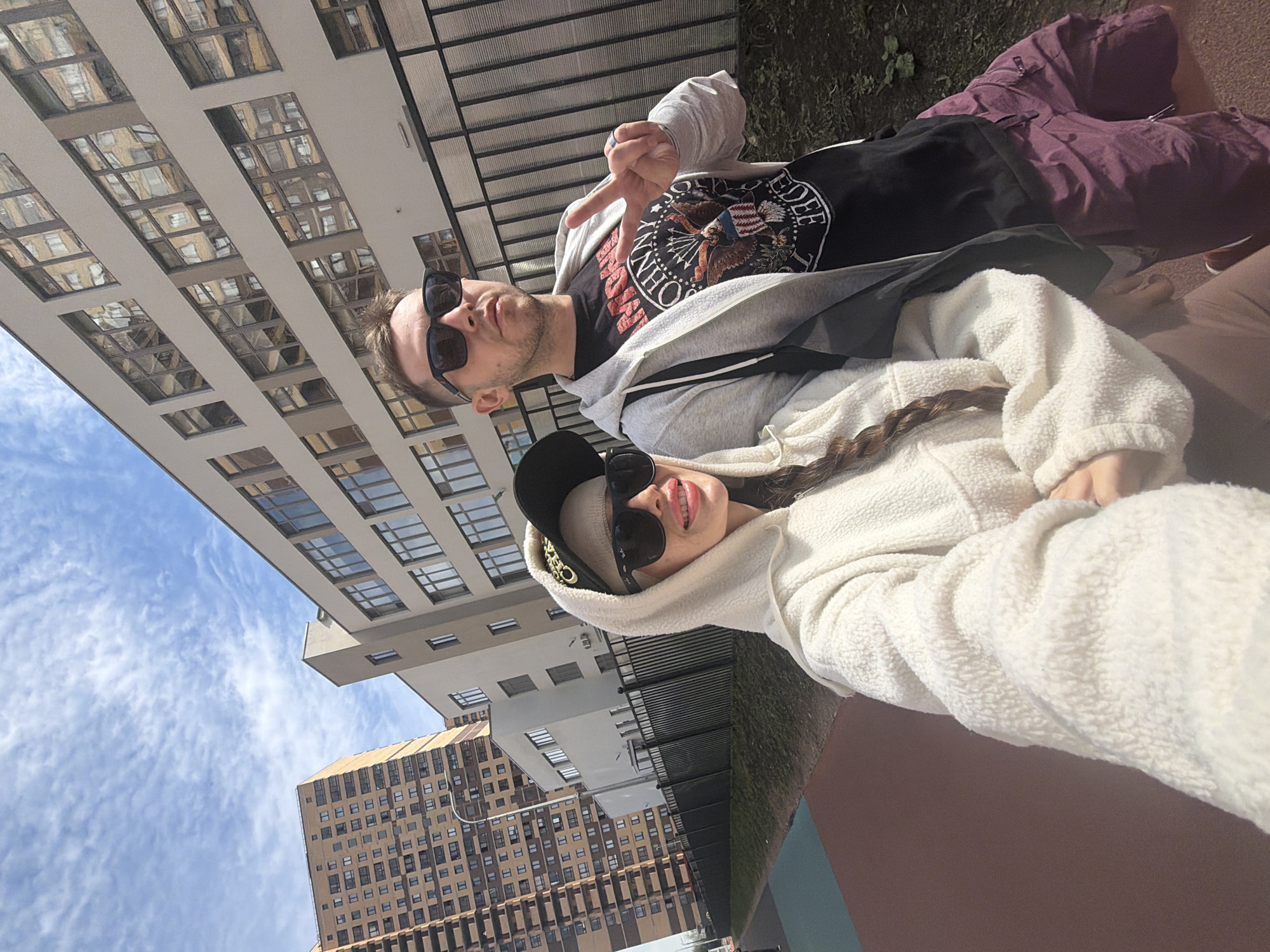 A man and woman stand outside on a sunny day in Saint Petersburg, Russia, framed against modern buildings and a blue sky with clouds. The man wears sunglasses, a gray hoodie, and a black t-shirt with a graphic. He extends two fingers in a peace sign. The woman wears sunglasses, a black baseball cap, and a fuzzy, hooded jacket. She has a braid visible from under the hood. The ground is a combination of grass and paving stones.
