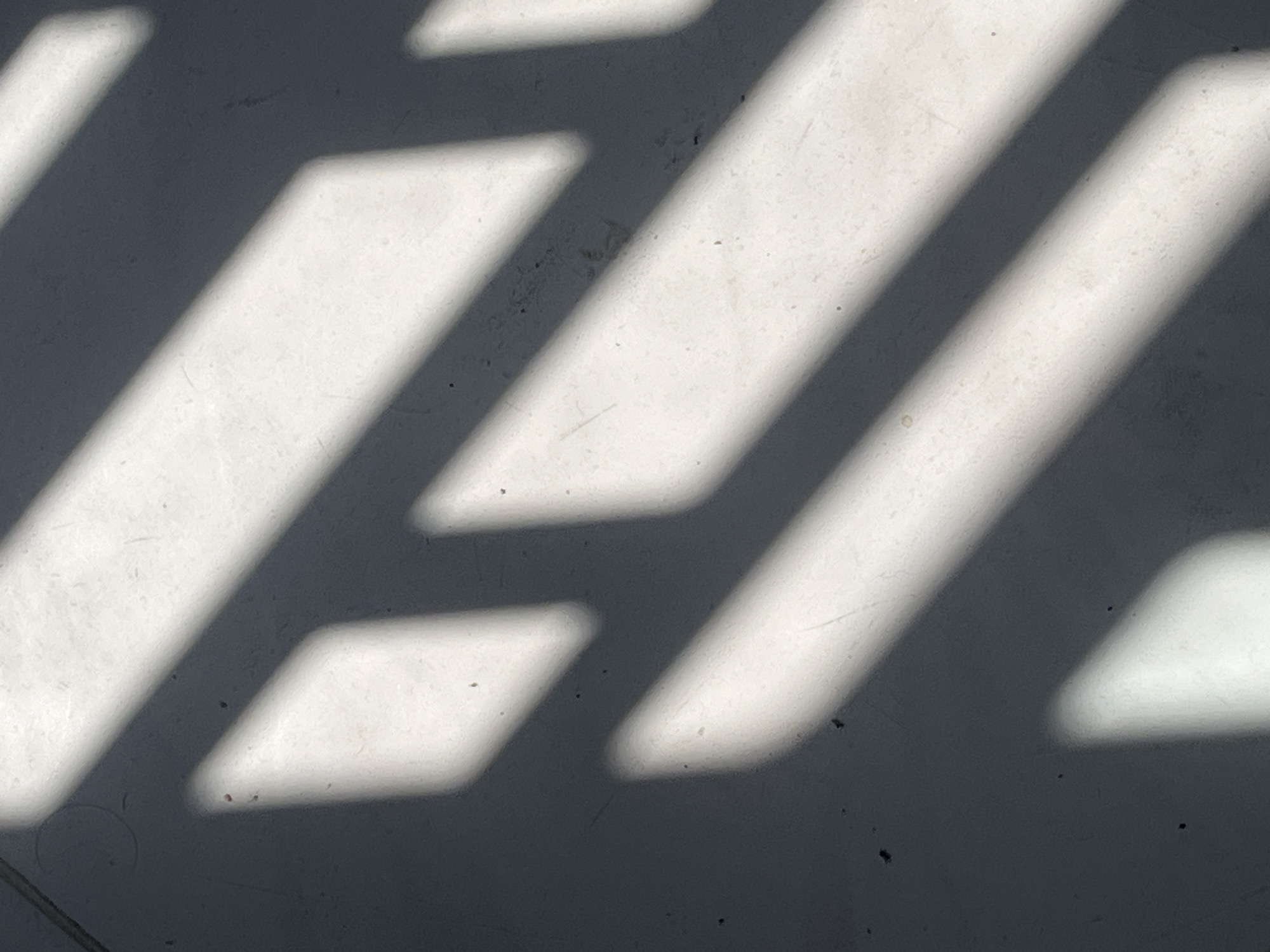 The image is an abstract composition of light and shadow on a light-colored, textured surface, likely a floor or wall. Parallel stripes of sunlight are cast diagonally across the frame, creating alternating bands of bright illumination and dark shadow. The shadows vary in width and intensity, with some appearing sharply defined and others softer. The surface itself shows subtle variations in tone and texture, with some small, dark specks visible within the lighter areas. The image does not contain any identifiable people, subjects, or actions, and therefore cannot be placed in Teluk Air, Indonesia, or any specific urban location.