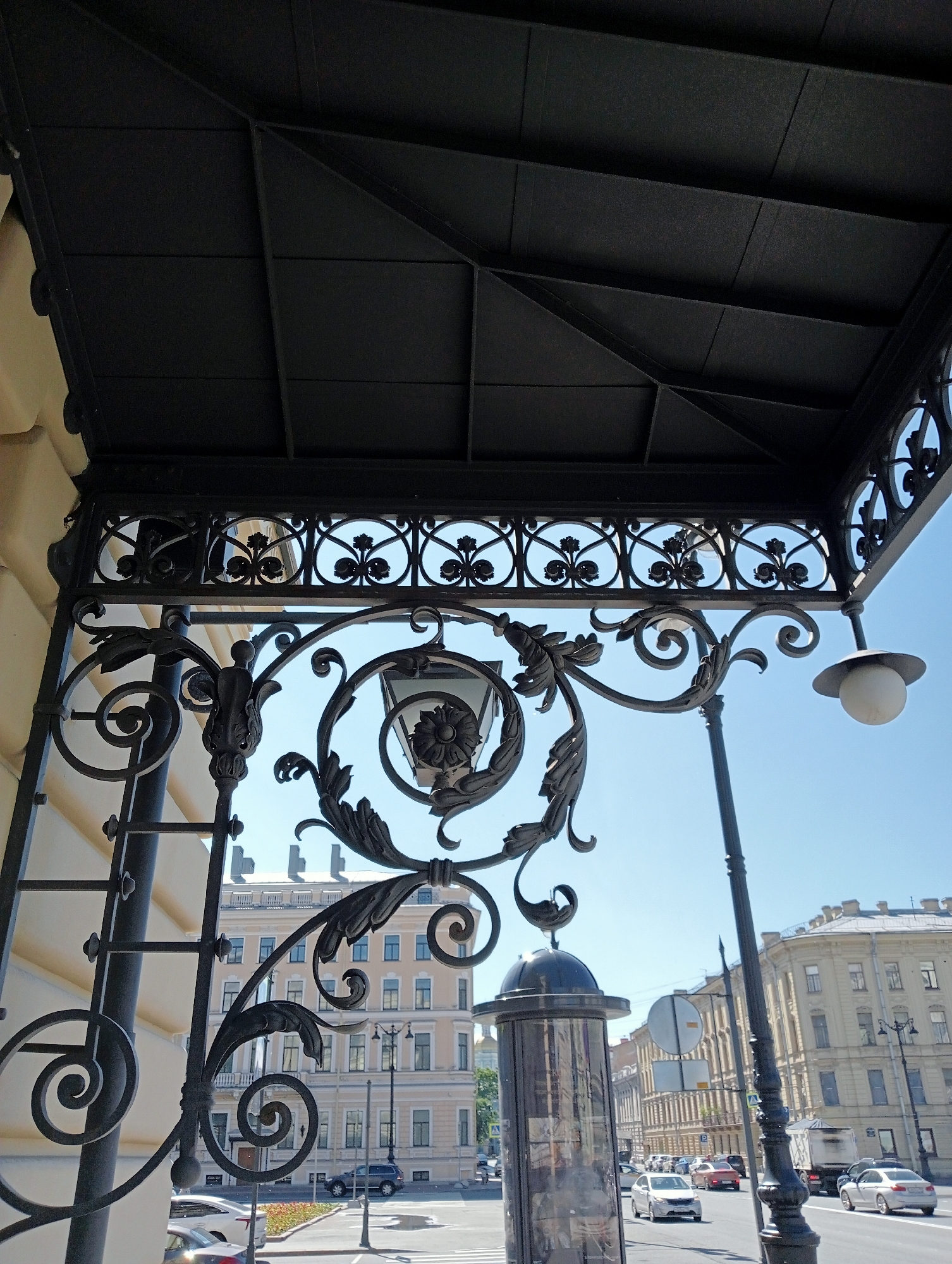 This is a low-angle, ground-level outdoor shot of an ornate black metal canopy structure with decorative scrollwork and floral motifs. A spherical white globe light fixture hangs from the right side of the canopy. Below and beyond the canopy, a street scene unfolds under a clear blue sky. Buildings with light-colored facades and numerous windows line the street. Cars, including a black SUV and a white sedan, are visible on the road, as are lampposts and traffic signals. A glass-enclosed structure, possibly a kiosk or bus stop shelter, stands near the foreground. The architecture suggests a location in Saint Petersburg, Russia, likely a historic district given the classical building styles and elaborate metalwork.