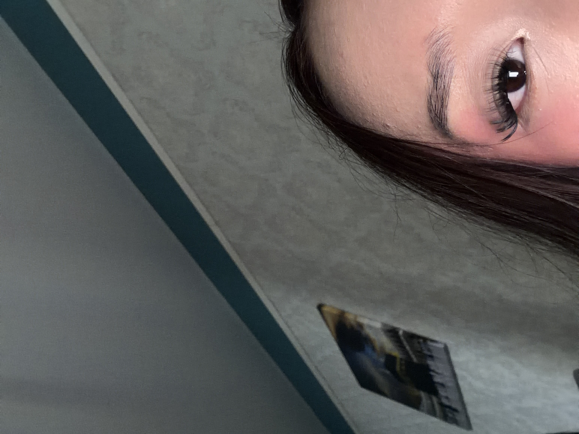 The image is a close-up shot of a person's eye and forehead. The person has dark hair, a visible eyebrow, and is wearing false eyelashes. There is pink blush on the cheek. The background appears to be a wall with a textured finish and a teal-colored border near the ceiling. A framed picture is visible on the wall to the left, which is abstract and appears to depict some sort of landscape. The lighting suggests an indoor setting, likely a room. There are no discernible details indicating the location in the city of Unknown, Russia.