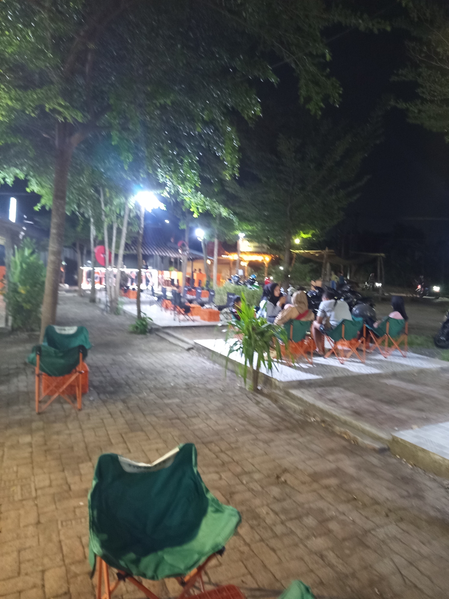 The image depicts an outdoor seating area at night, likely a cafe or restaurant, in Becirongengor, Indonesia. Several people are seated in orange folding chairs covered with green fabric, engaged in conversation. In the foreground, a similar chair is empty. The area is paved with cobblestones, and trees line the walkway, their leaves illuminated by artificial lighting. In the background, more seating arrangements, tables, and motorcycles are visible, suggesting a lively social gathering place. The overall atmosphere is relaxed and casual, with the scene illuminated by a combination of ambient lighting and streetlights.
