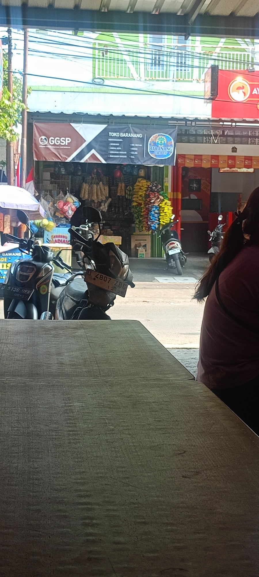 The image depicts a street scene in Mekar Rahayu, Indonesia. In the foreground, a weathered wooden table occupies the lower portion of the frame. Behind the table, two motorcycles are parked. The motorcycle on the left is black and blue, with a license plate reading "D 5136 UER". The motorcycle on the right is black, with a license plate reading "D 4807 ZBG".
In the background, a store or shop is visible, identified by banners. One banner reads "GGSP". Another banner above the shop entrance displays "TOKO BARANANG" with various items listed below, including "BAWANG MERAH", "BAWANG PUTIH", "ANEKA KOPI", and "MAKANAN RINGAN". A circular logo with text "BARANANG GROUP" is also present on this banner. The shop's facade features displays of goods such as what appear to be dried snacks or food items hanging in bunches, and colorful decorative items. A red sign with a white circular logo is visible to the right of the shop. A person with dark hair, wearing a maroon shirt, is partially visible on the right side of the image, facing away from the camera. The environment appears to be a commercial area with overhead wires and some greenery on the left. The lighting suggests it is daytime, with the sun casting shadows.