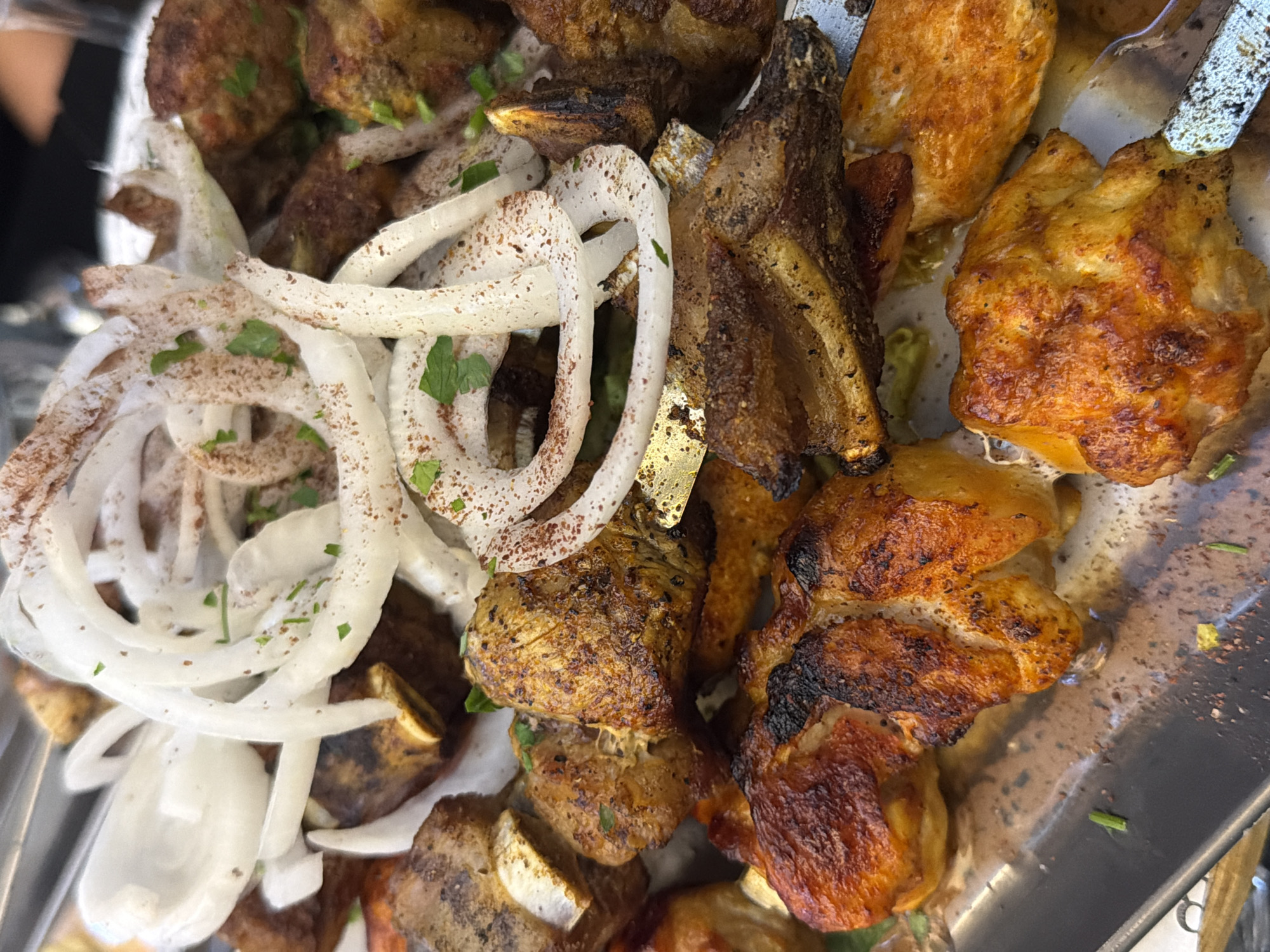 A high-angle close-up shot shows a metal tray filled with cooked meats and onion rings. The meats consist of various cuts, some heavily charred, suggesting grilling or roasting. Sliced white onion rings are piled on top, sprinkled with a light green herb. The location appears to be Rishon LeZion, Israel, based on identifying marks.