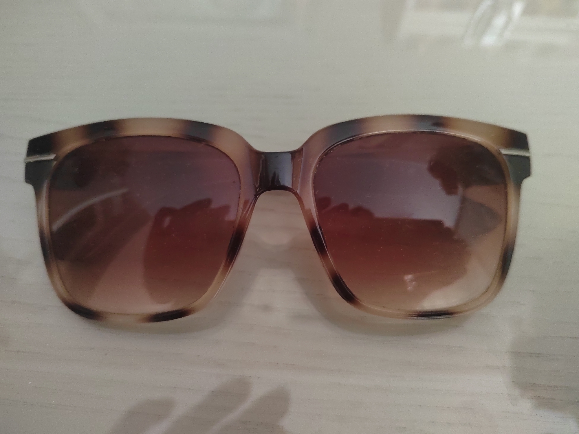 A pair of tortoise shell framed sunglasses with gradient brown lenses are positioned flat on a light-colored, horizontally-lined surface. The sunglasses are centered in the frame, with the left lens showing a reflection of a light source. The background is out of focus and appears to be a wall or similar surface. There are no people present and the scene is static. The location is not specified.