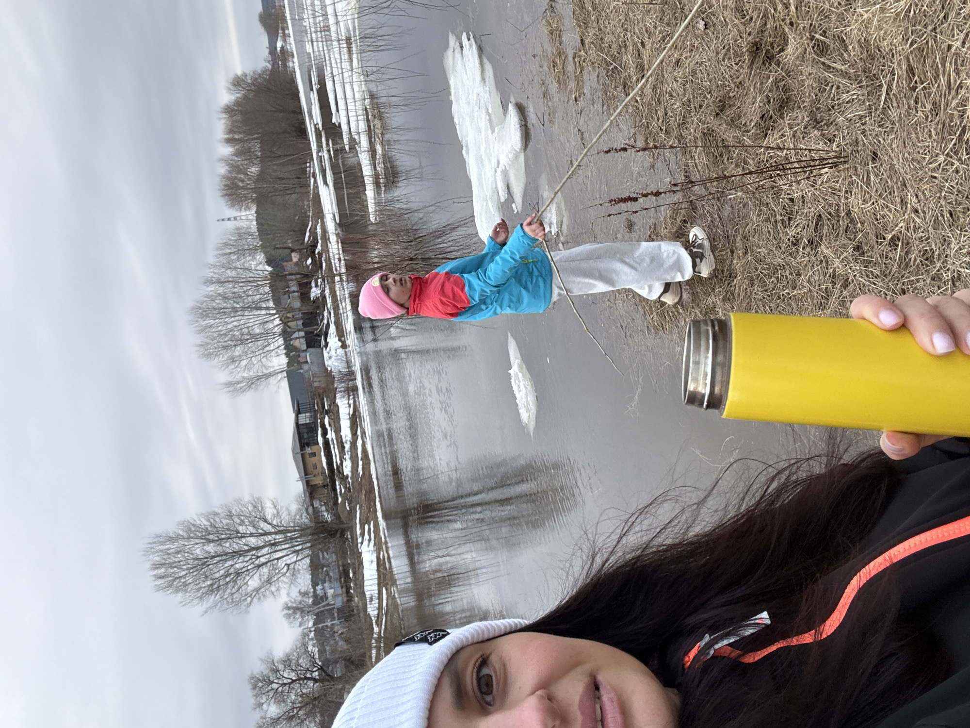 A selfie shows a woman holding a yellow water bottle. In the background, another person is near a body of water covered with ice. The location appears to be in or near the city of Chaikovsky, Russia.
